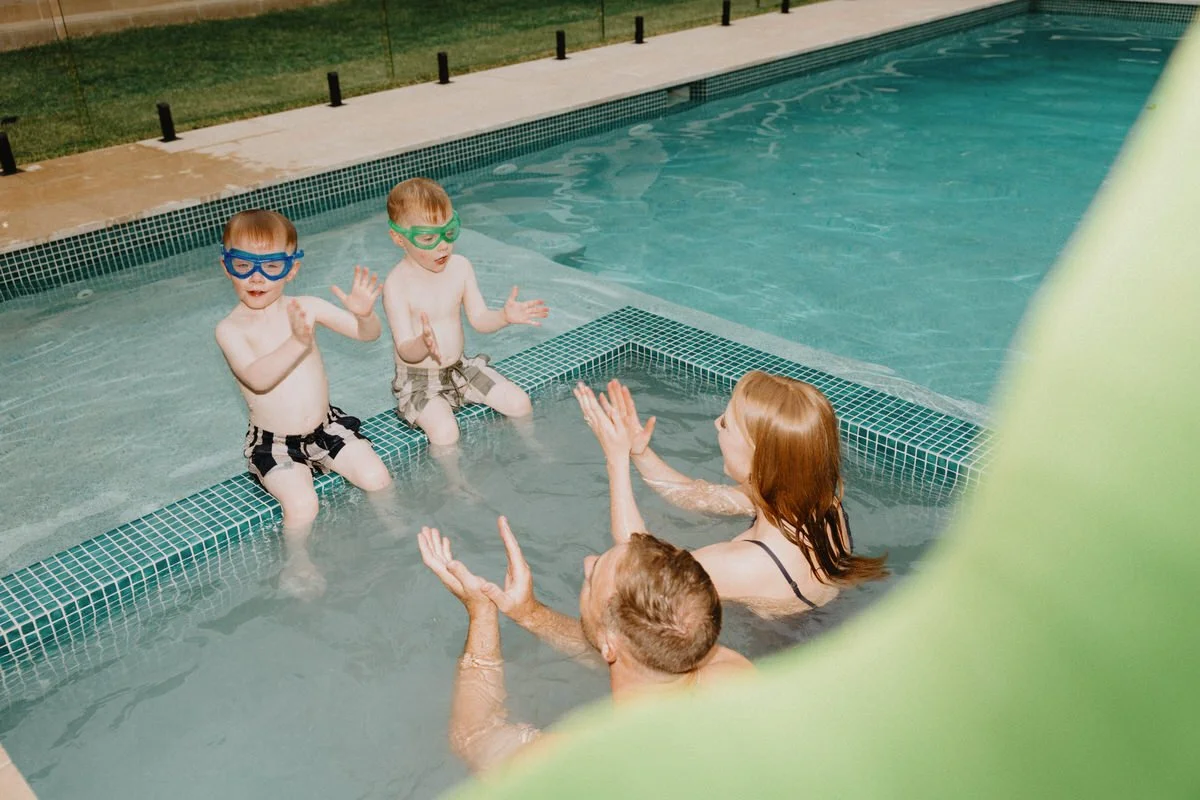Two young boys and two adults enjoying a swim in a pool. The boys are wearing swim trunks and goggles, while the woman and man are in the pool with the children, engaging in a playful activity in the water.