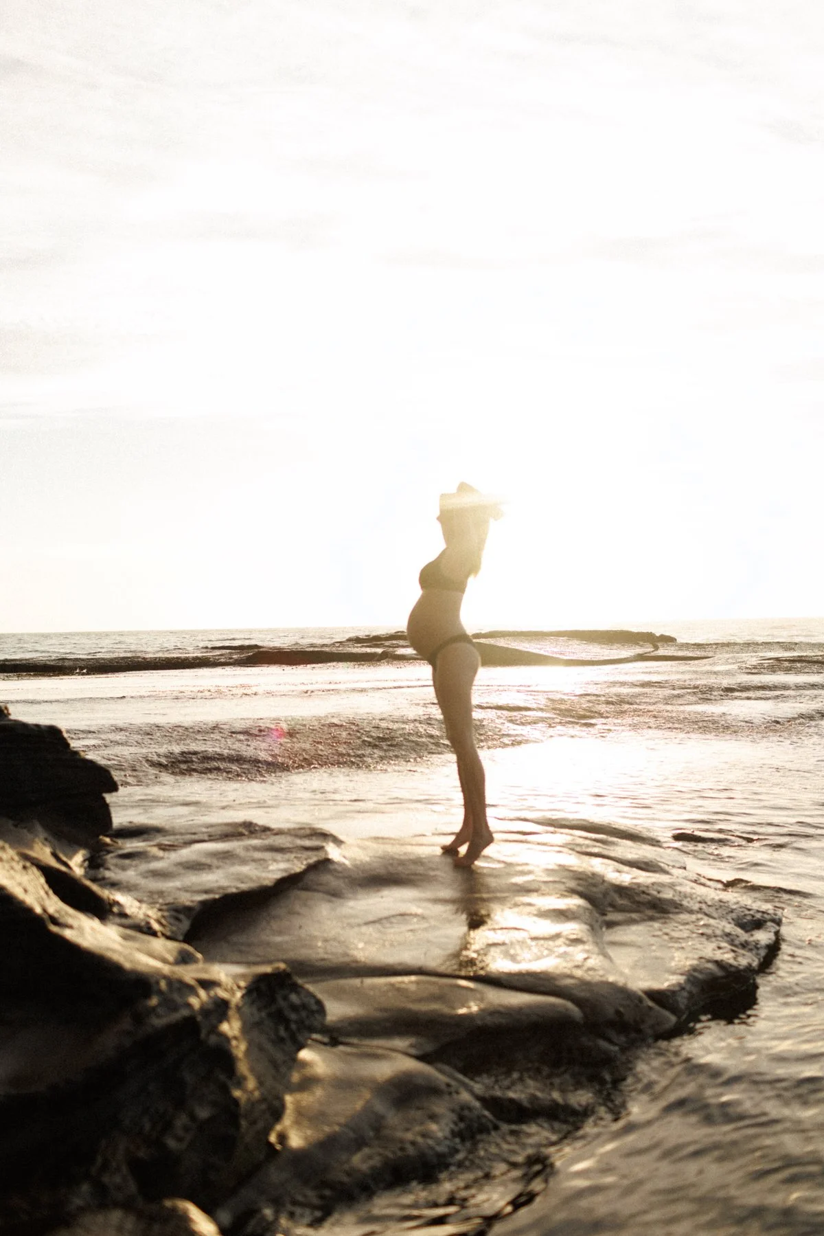 A woman standing on a rock at the beach during sunset, wearing a swimsuit and a hat, with the ocean in the background.