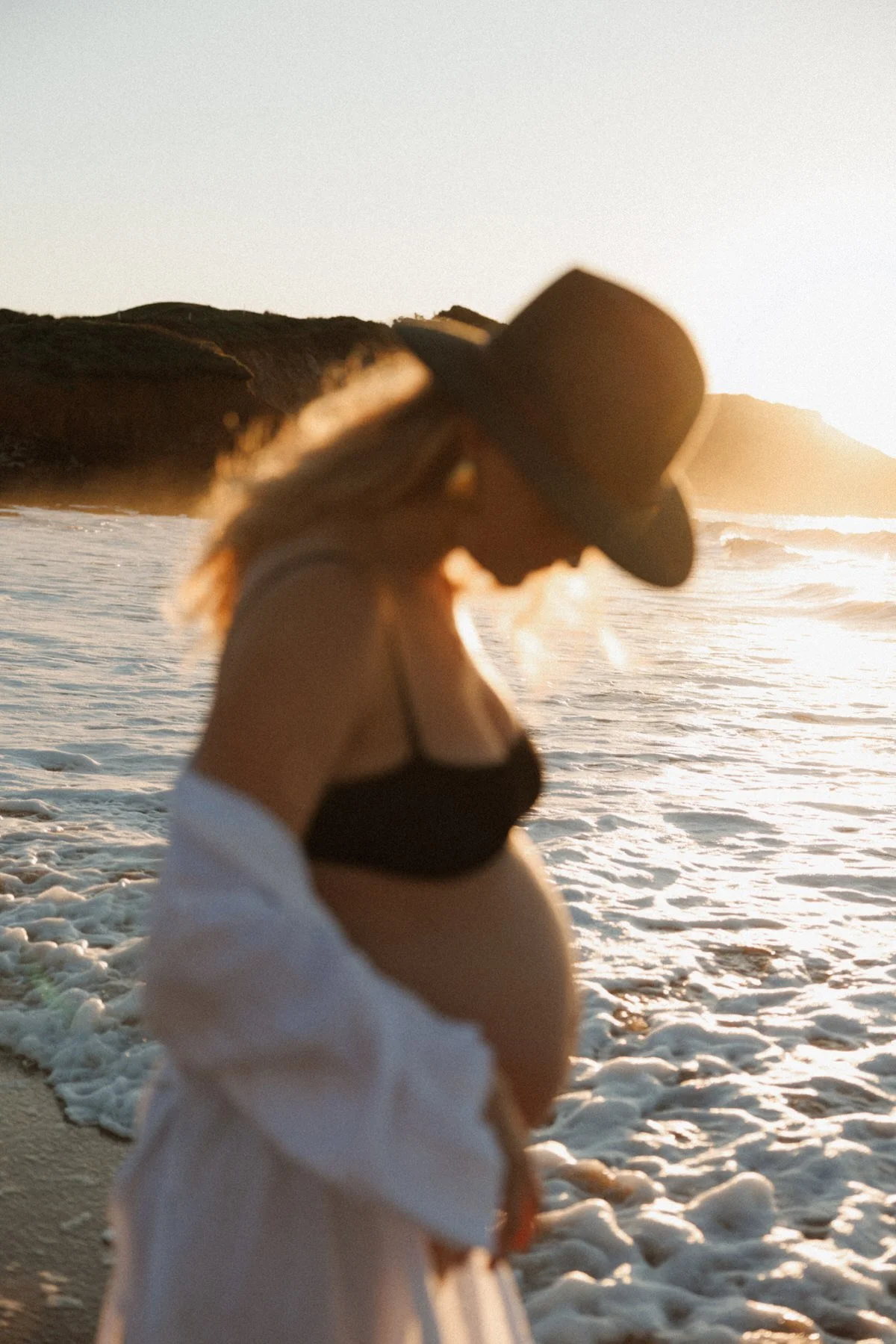 A woman in a black bikini top and a white cover-up with a wide-brimmed hat, standing on a beach at sunset, with waves crashing behind her and head slightly bowed.
