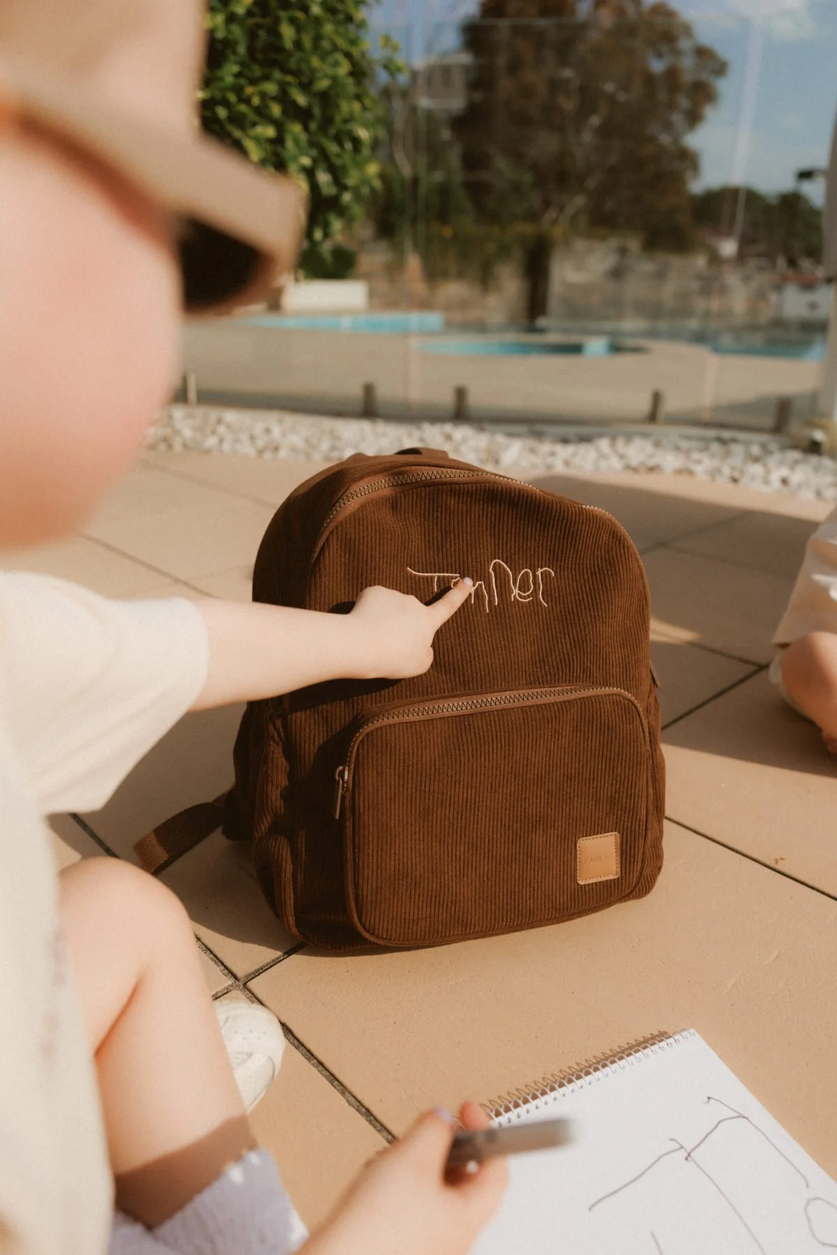 Child pointing at a brown backpack with the name "Tomler" embroidered on it, sitting outside on a tiled surface with a school notebook, and a piece of paper with handwriting. In the background, there are trees, a fence, and a swimming pool.