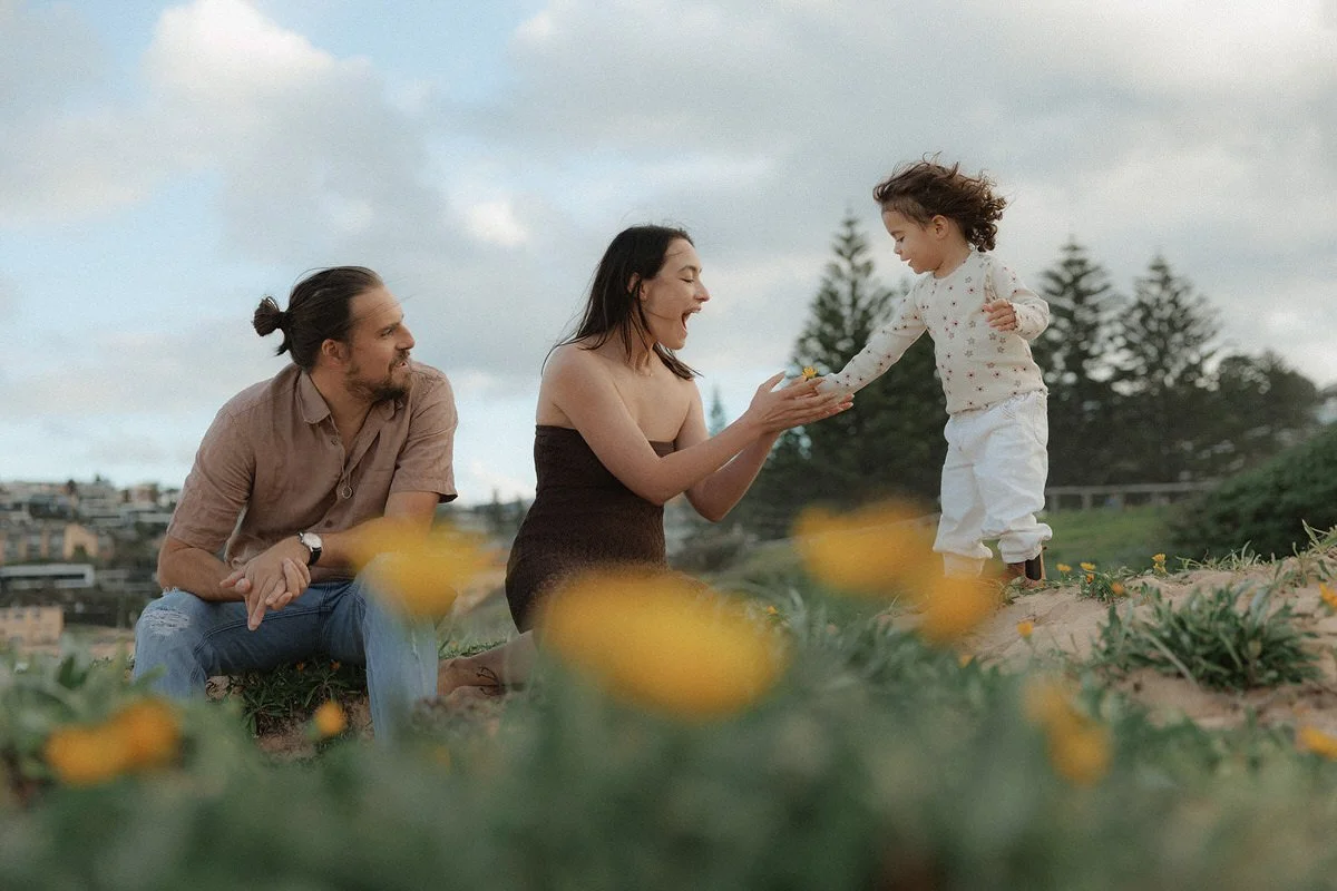 A woman and a young girl exchanging a flower outside with a man smiling nearby, in a grassy area with yellow flowers and trees in the background.