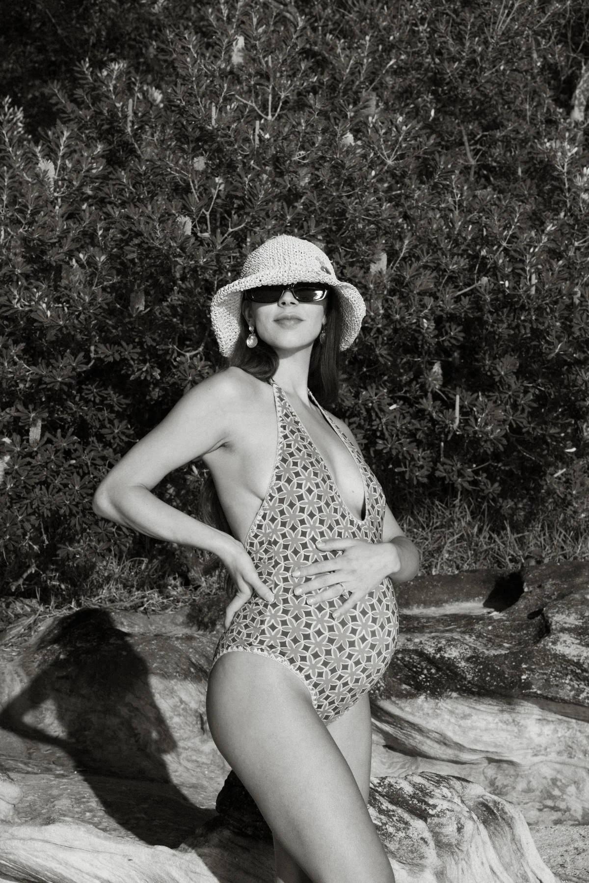 A woman in a swimsuit, hat, and sunglasses standing on a rock in front of dense foliage, posing with one hand on her hip and the other resting on her stomach, in a black and white photo.