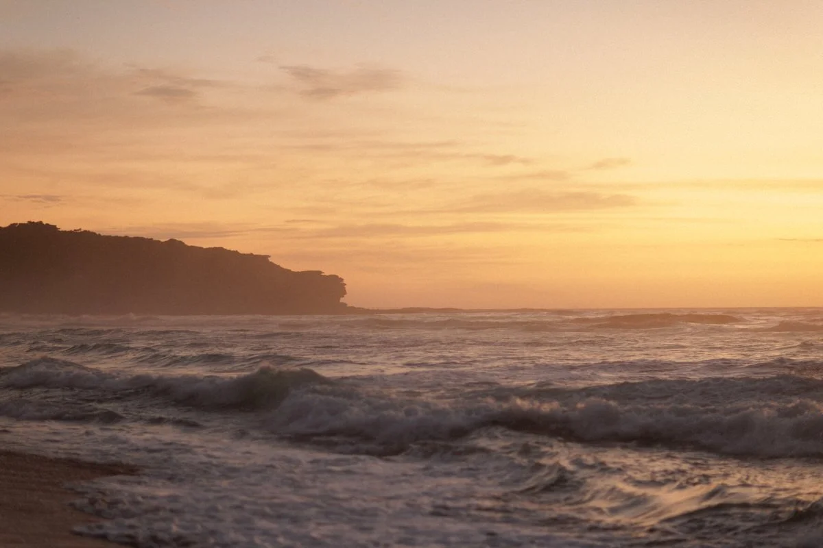 Sunset over the ocean with waves crashing on the shore and a silhouette of a distant cliff.