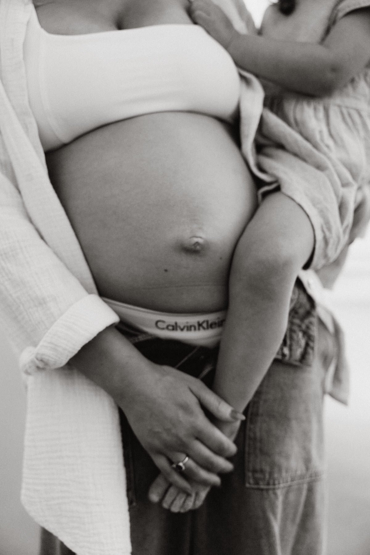 A close-up of a person with a pregnant belly, wearing Calvin Klein underwear, with another person beside them gently touching their belly, their hands and part of their torso visible.