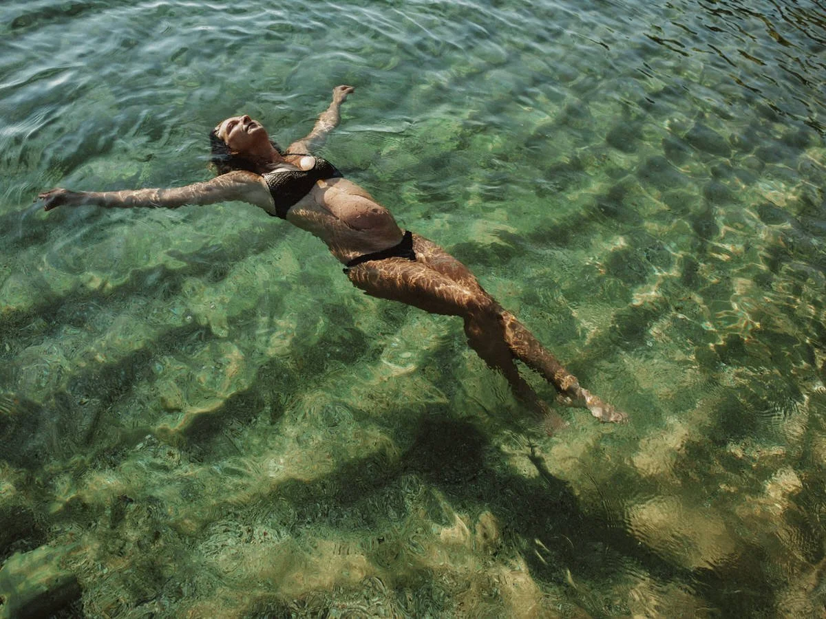 Woman floating peacefully in clear green water, arms and legs extended, wearing a black swimsuit.