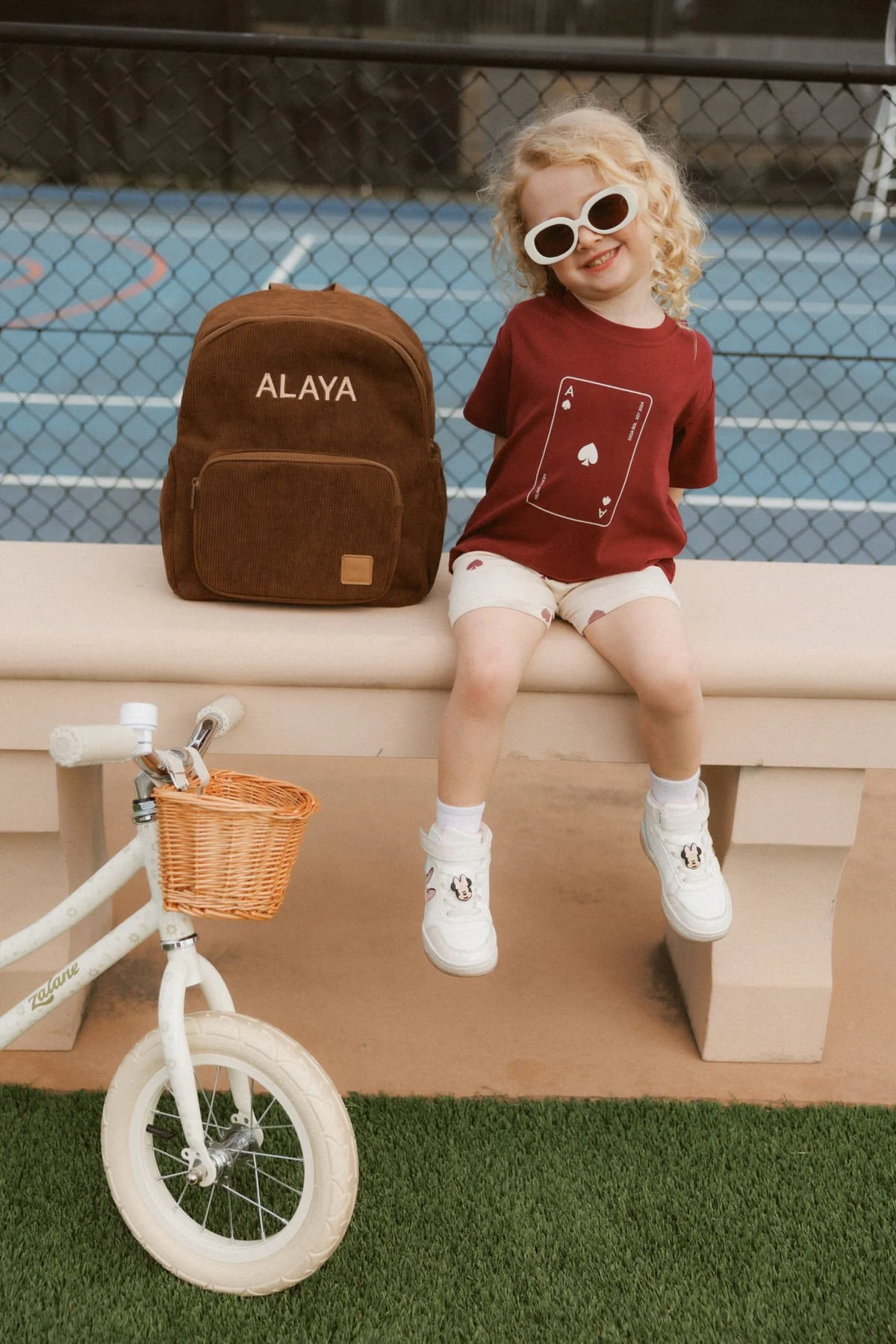A young girl with curly blonde hair wearing sunglasses, a red t-shirt with a playing card graphic, white shorts, and sneakers with Minnie Mouse patches, sitting on a bench with her legs dangling. Next to her is a brown backpack with the name 'ALAYA' 