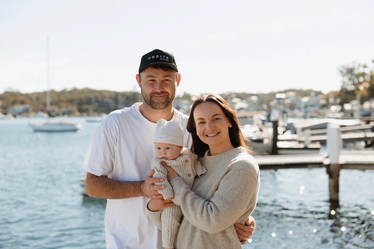 A smiling family of three, a man, woman, and baby, standing by a marina with boats and a dock in the background on a sunny day.