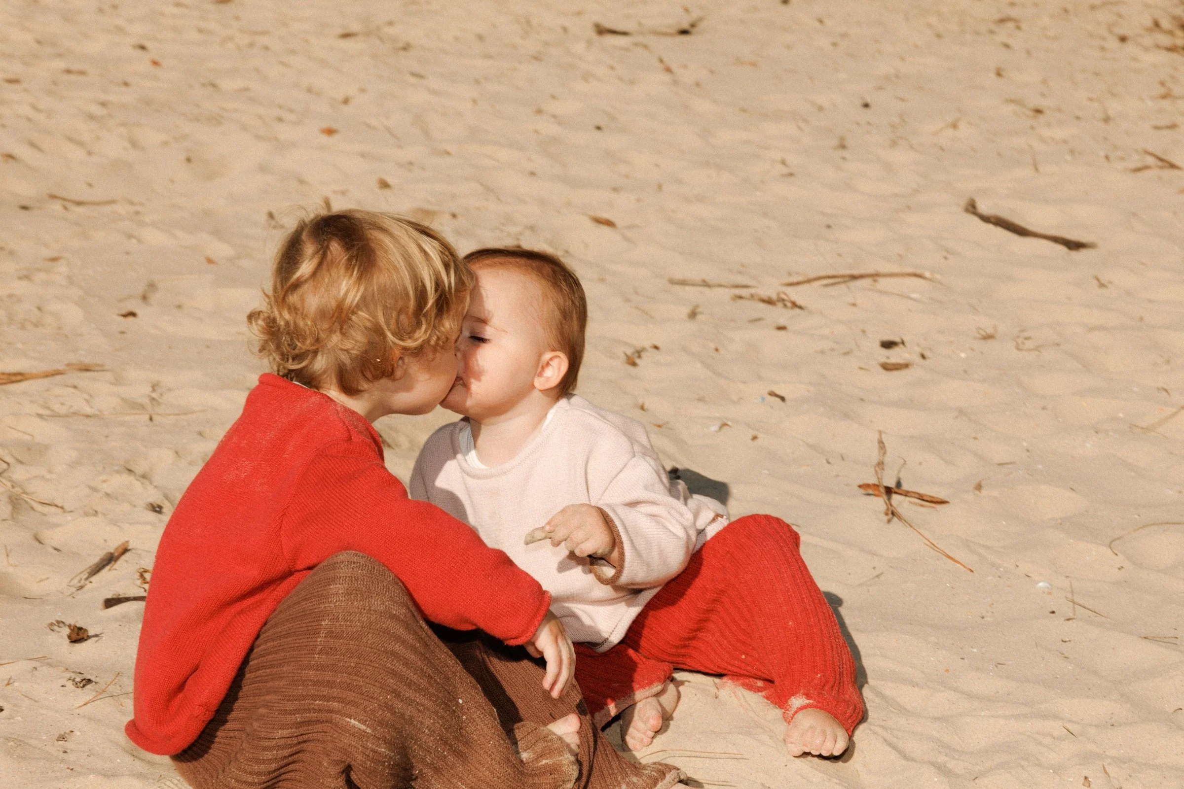 Two young siblings sharing a kiss while sitting together on the sand.