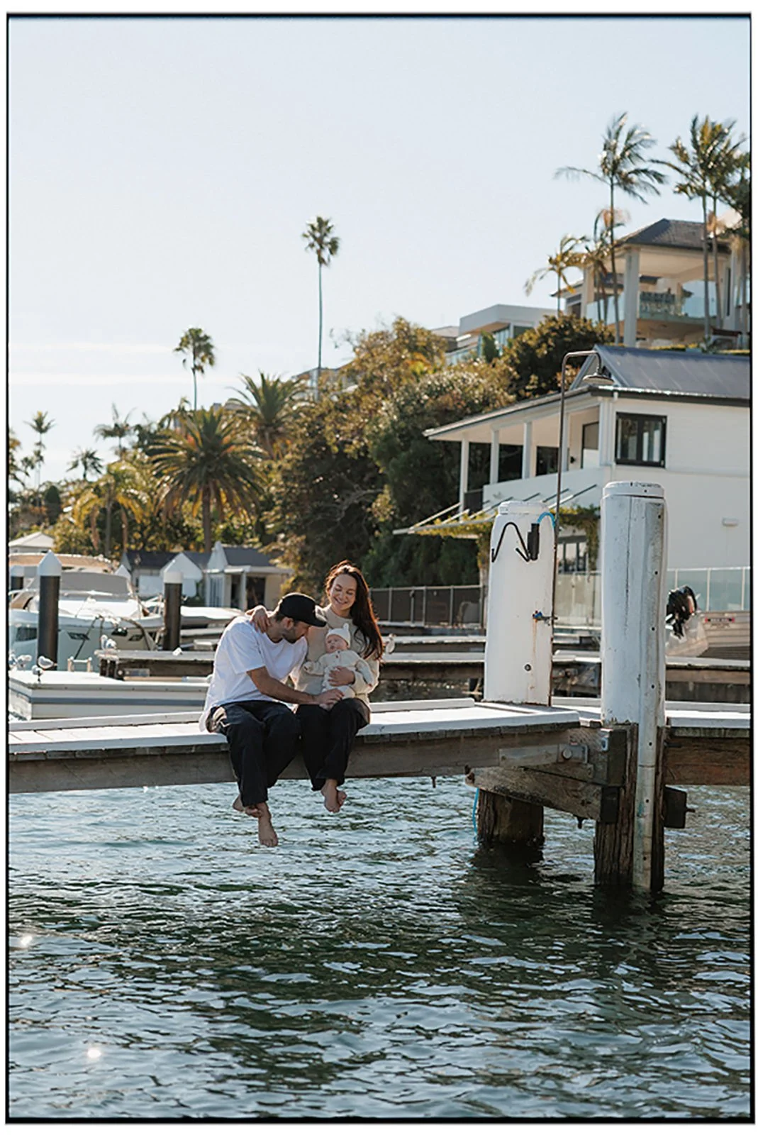 A family sitting on a dock by the water, holding a baby, with a house, palm trees, and boats in the background on a sunny day.