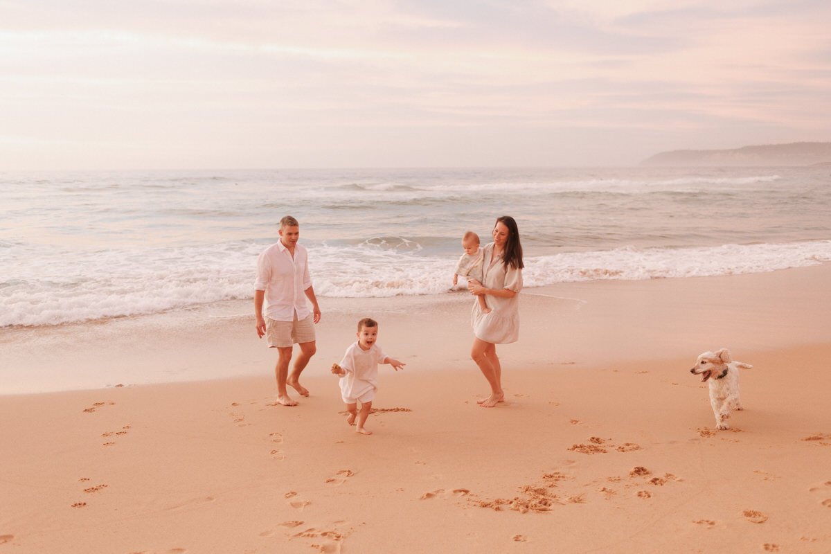 Family with two children and a dog walking on the beach near the shoreline during sunset.
