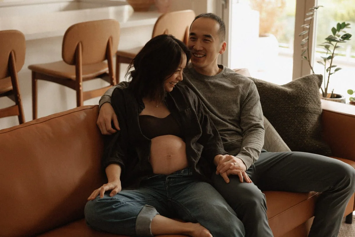 A pregnant woman and a man sitting on a couch in a living room, smiling and holding hands.