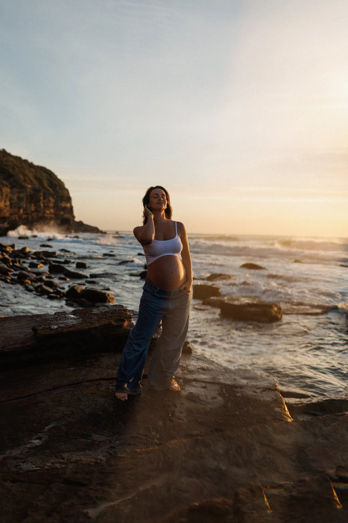 A pregnant woman standing on a rocky beach during sunset, wearing a white tank top and jeans, with her hand touching her ear and a serene expression.