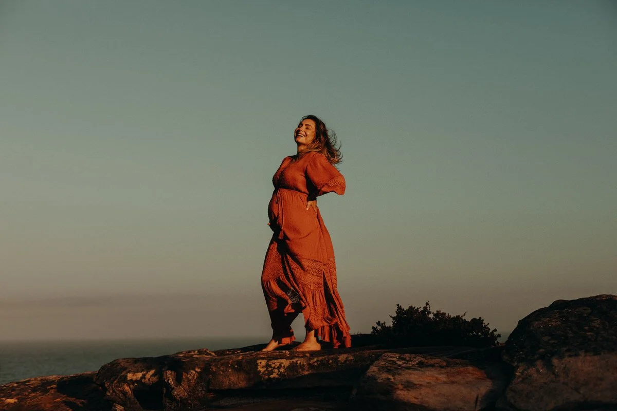 A woman standing barefoot on rocks outdoors during sunset, wearing a long orange dress, smiling and looking up.