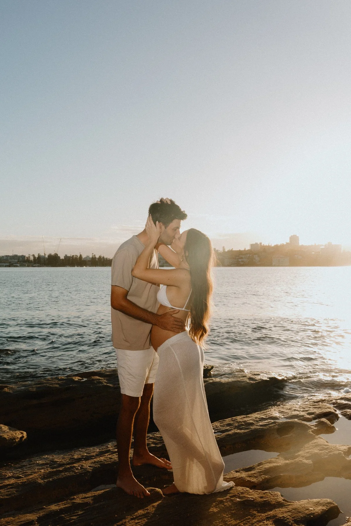 A couple kissing on a rocky beach at sunset, with water and city skyline in the background.