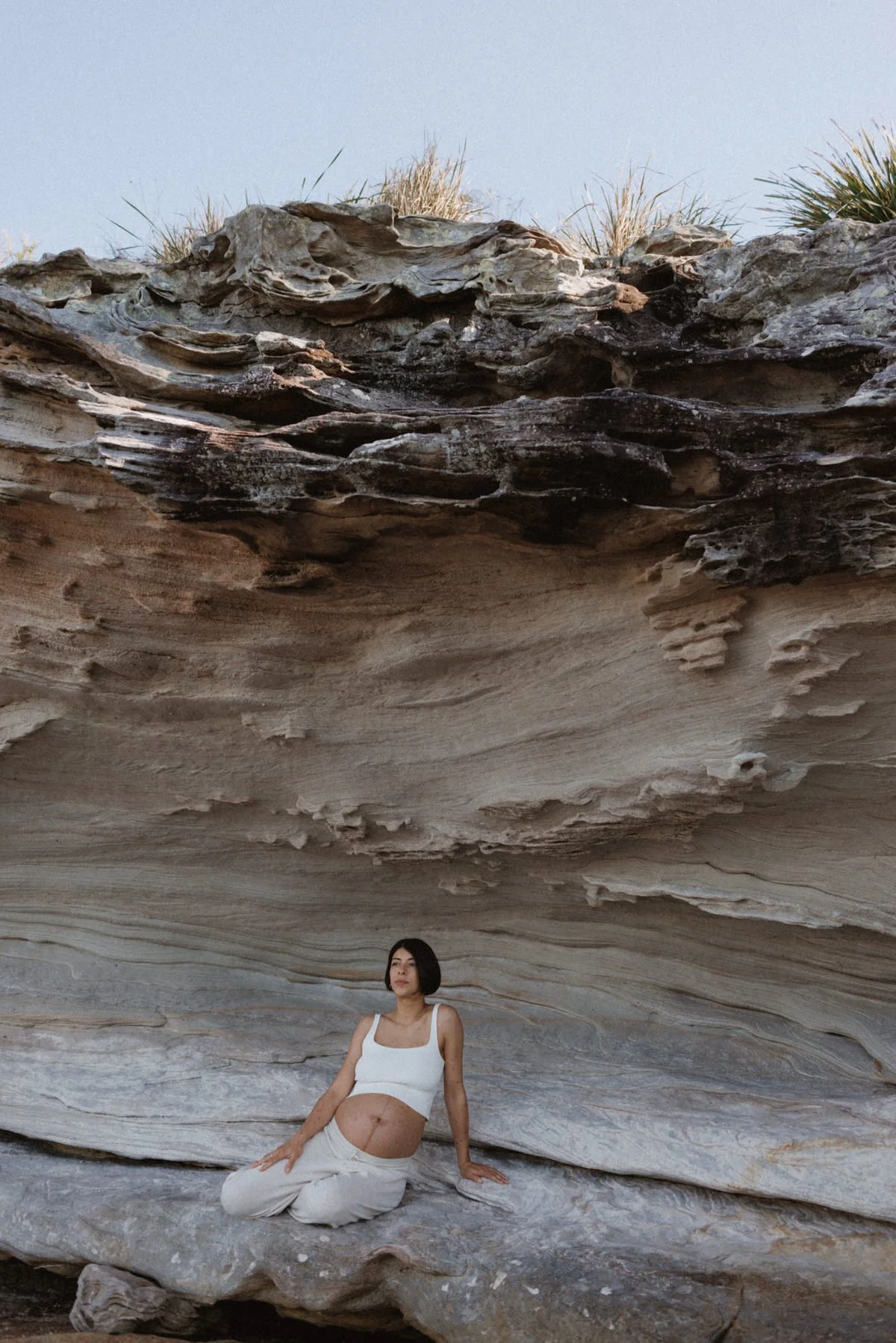 A woman in white clothing sitting on a large, smooth rock formation with layered, striated rock surfaces beneath a large overhanging rock and some grass at the top.