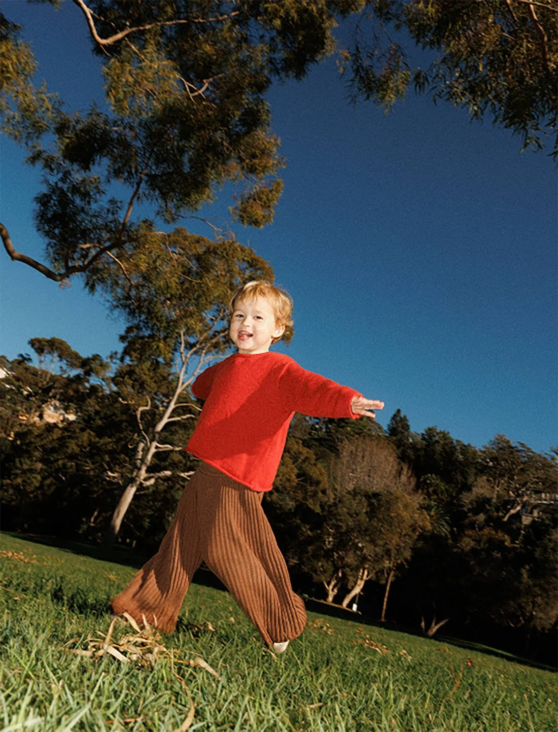 A young child with blonde hair wearing a red sweater and brown pants playing outdoors on a grassy field with trees and a clear blue sky in the background.