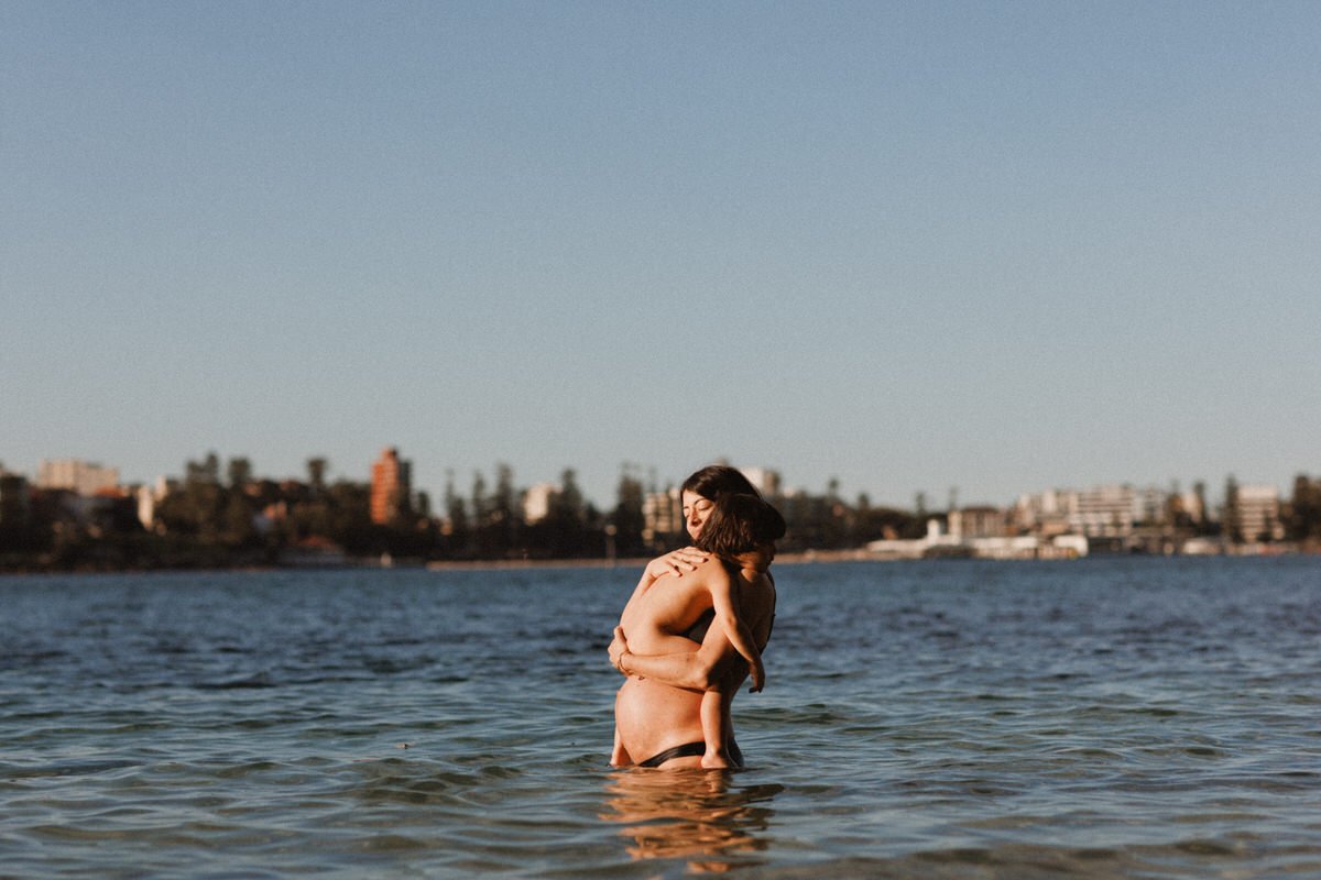 A woman holding a child while standing in the water at the beach during sunset with a city skyline in the background.