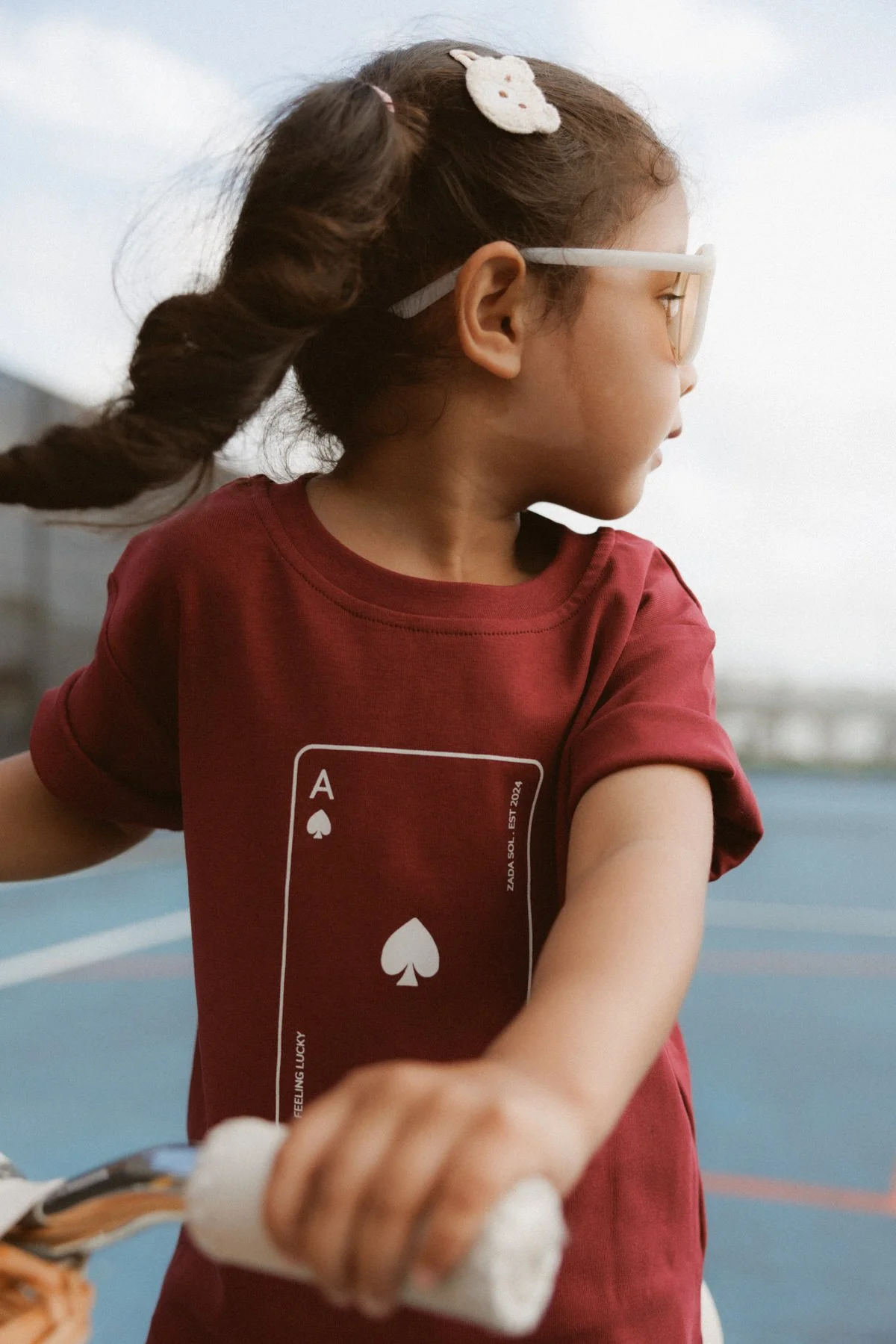 A young girl with brown hair tied in a braid, wearing white glasses and a red T-shirt with a playing card graphic, is looking to her side while holding a white object in her hand outdoors.