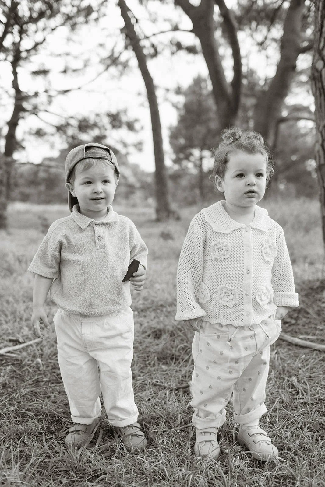 Two young children, a boy and a girl, standing outdoors on grass with trees in the background. The boy is wearing a cap, polo shirt, and pants, holding an object in his hand. The girl is wearing a textured sweater with floral patterns and pants. Both