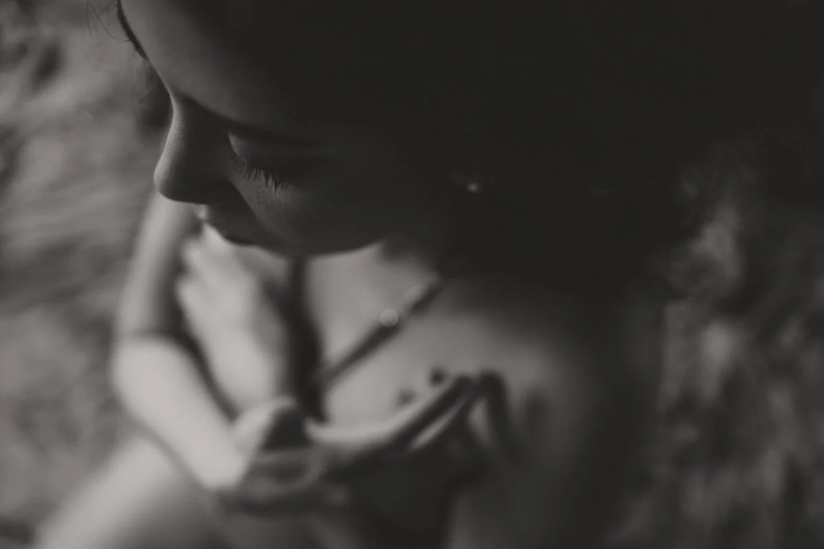 Black and white close-up of a young girl looking down, with her arms crossed, wearing glasses and a chain necklace, with blurred background.