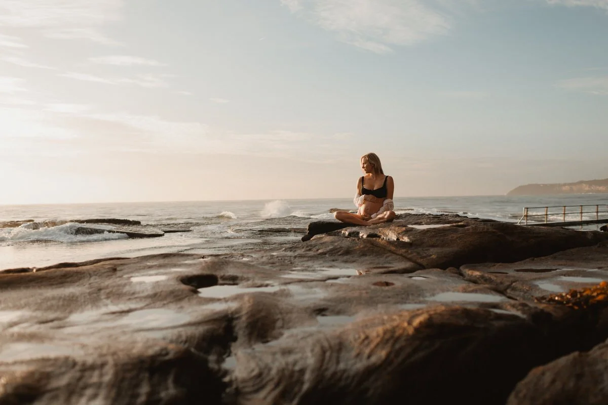 A pregnant woman sitting on rocks near the ocean during sunset, looking out at the water.