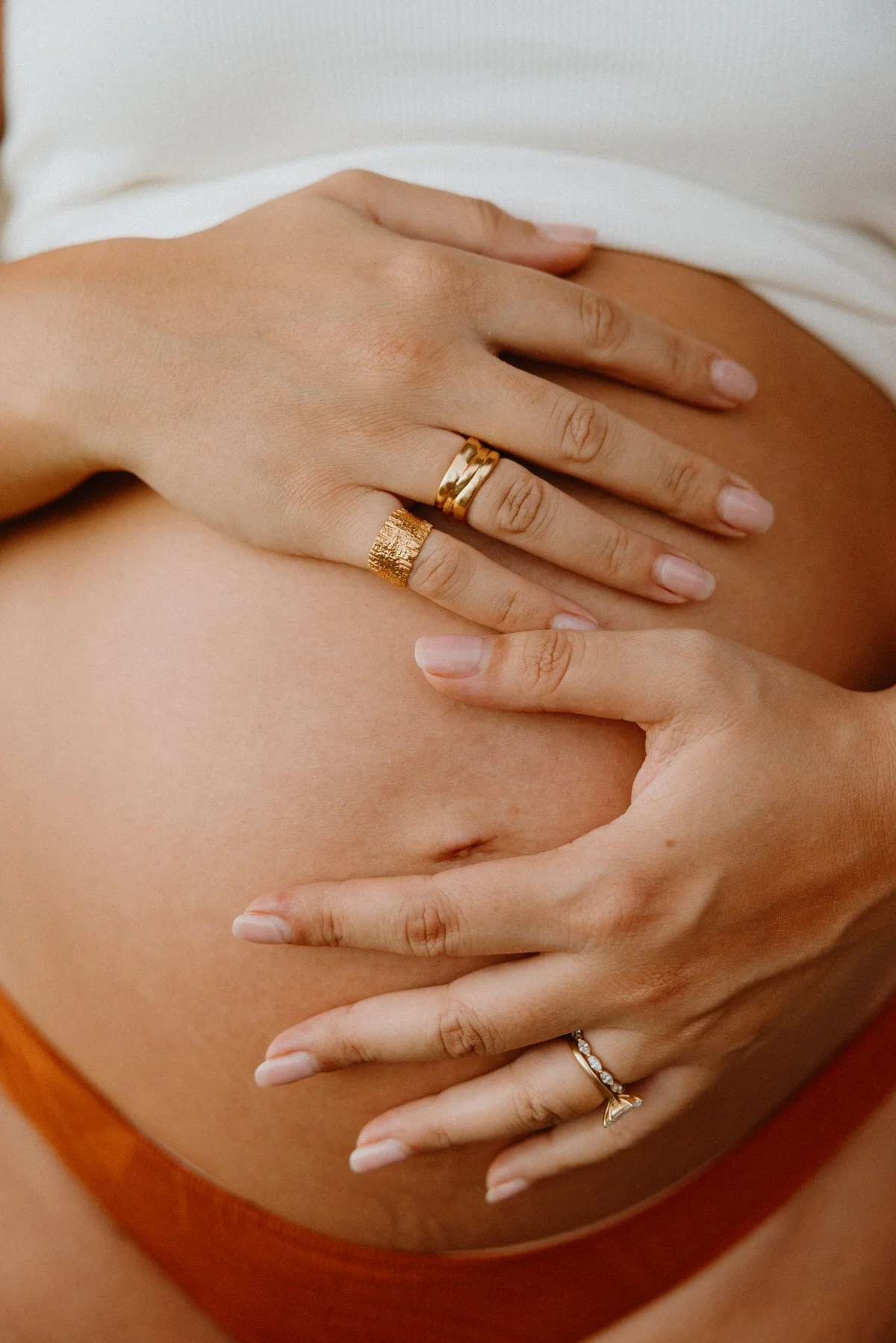 Close-up of a person's hands gently resting on their bare stomach, adorned with gold and silver rings.