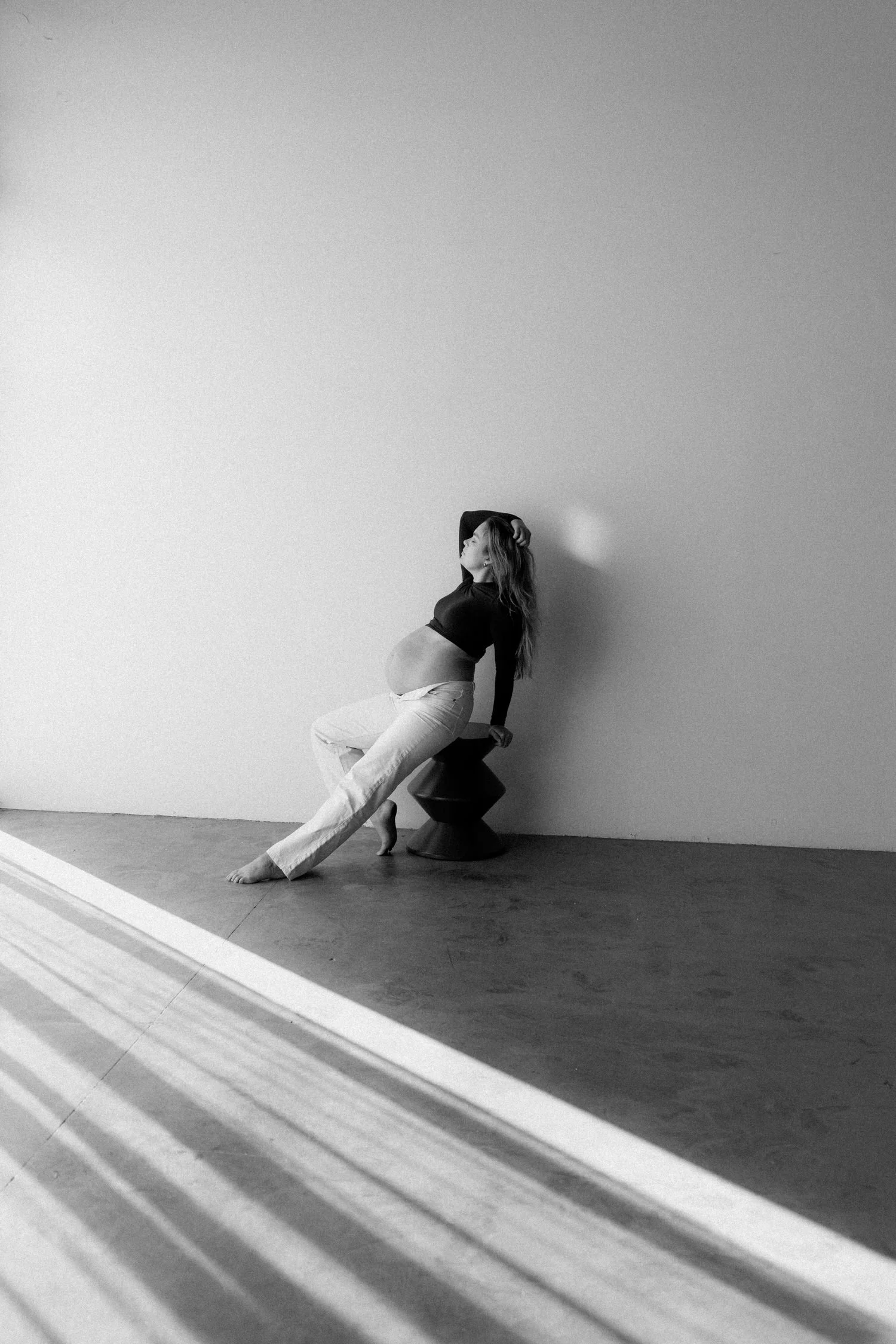 Black and white photograph of a woman with long hair sitting on a stool against a plain wall. She is dressed in a dark top and light pants, with her head tilted back and one arm raised to touch her hair. The photo is taken through a window with horiz