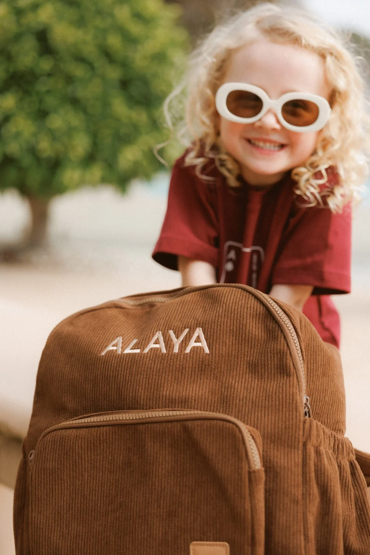 A young girl with curly blonde hair wearing white sunglasses and a red shirt, smiling behind a brown bag with the name "ALAYA" embroidered on it, outdoors with a green tree in the background.