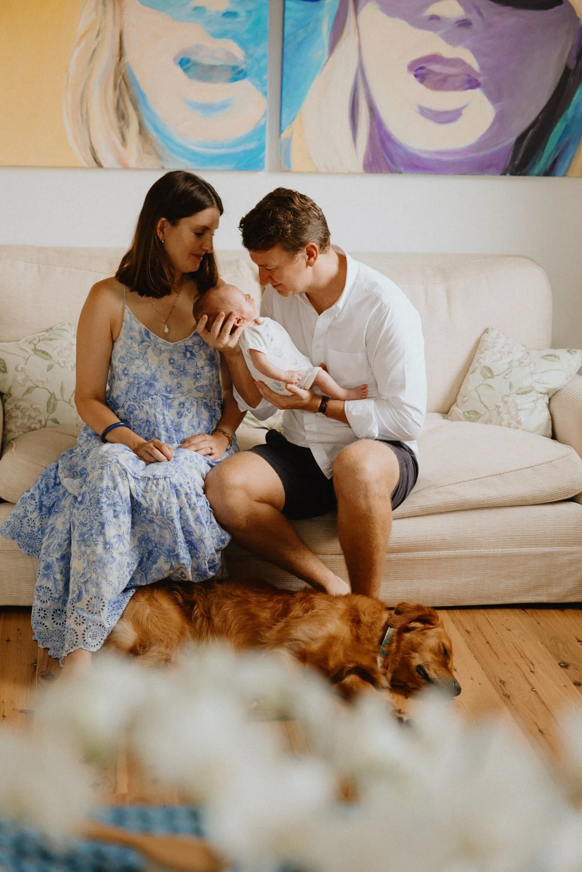 A family sitting on a beige couch with a baby, woman in a blue dress and man in a white shirt. Two golden retrievers are lying on the wooden floor in front of the couch. Two colorful abstract portraits hang on the wall behind them.
