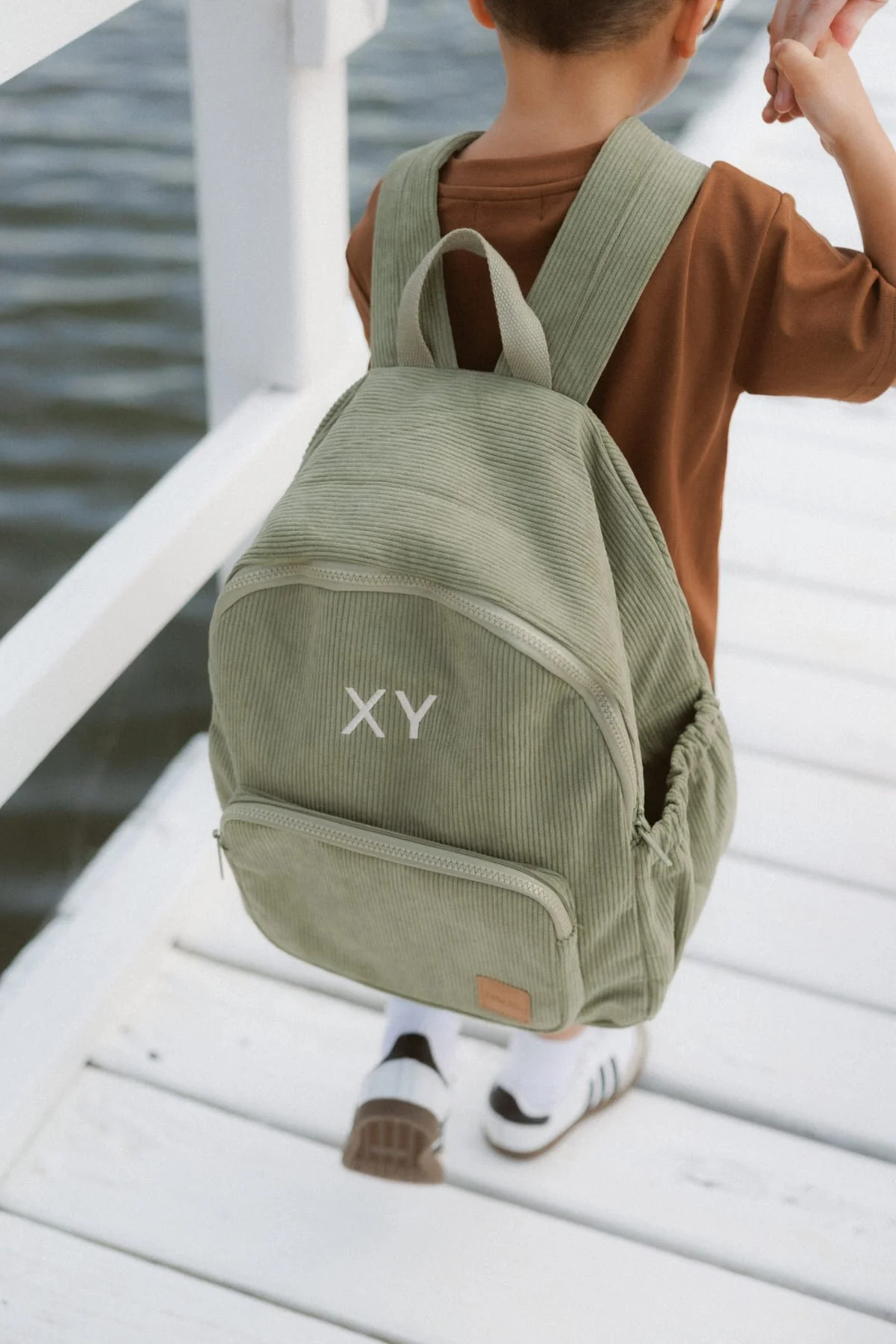 Child with a beige backpack featuring 'XY' on the front pocket, standing on a white bridge with water below.