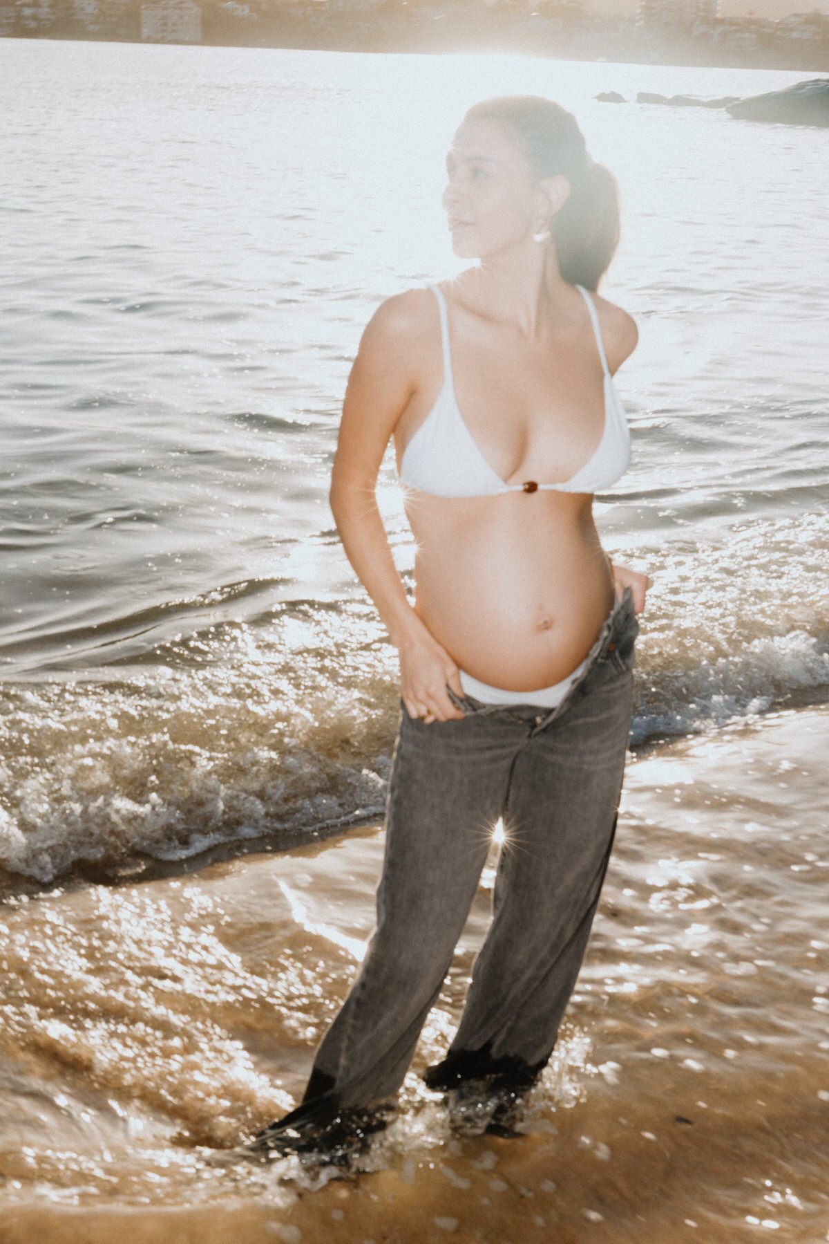 A pregnant woman in a white bikini top and loose jeans standing in water at the beach, with her hand in her pocket, facing the right, and smiling.