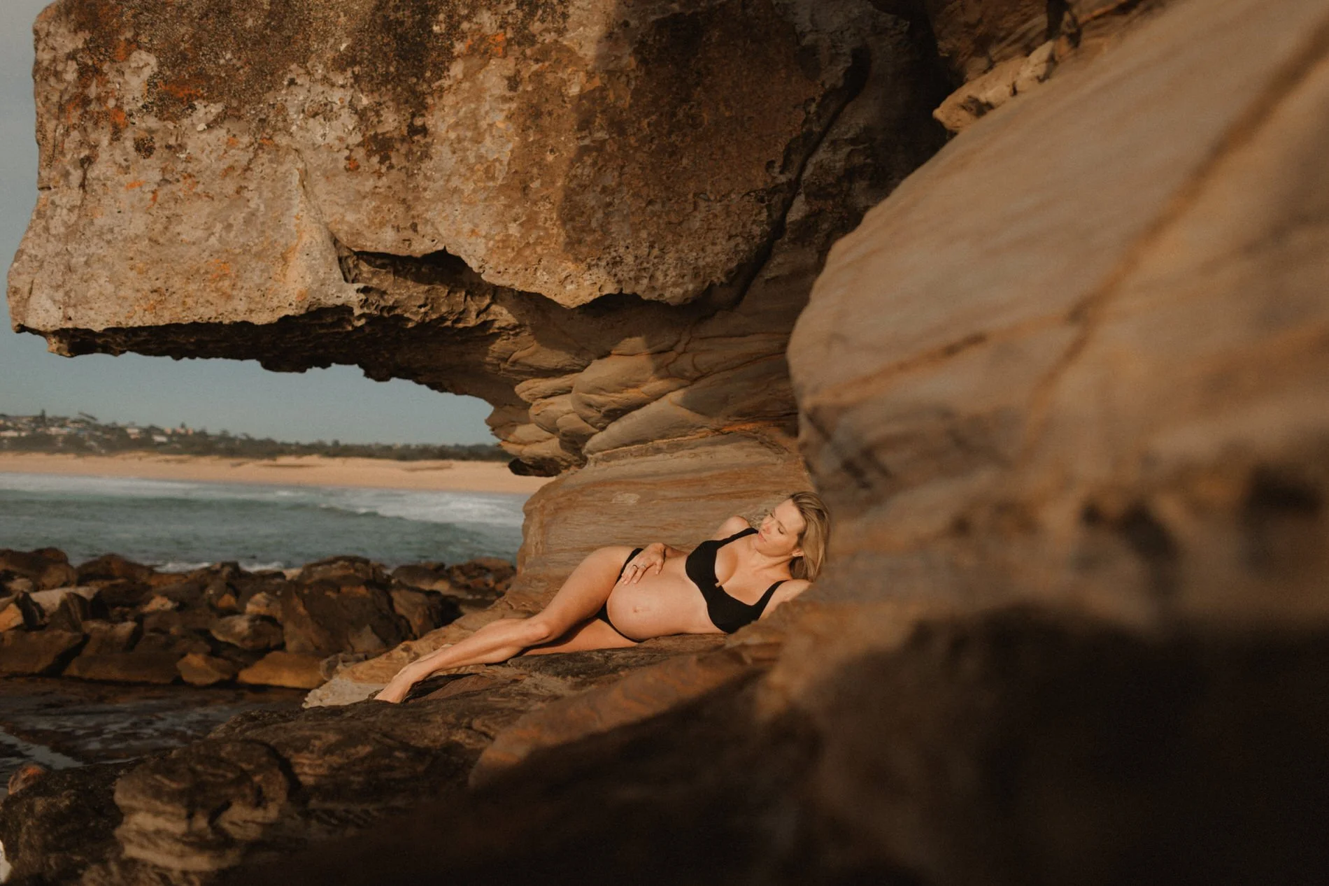 A woman in a black swimsuit lying on a rock formation near the beach, with large sandstone rocks over her and the ocean in the background during sunset.