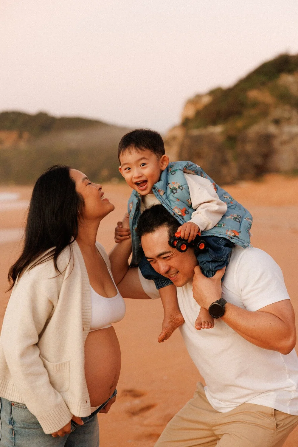 Pregnant mother smiling beside her partner, with their laughing toddler on his shoulders at the beach.