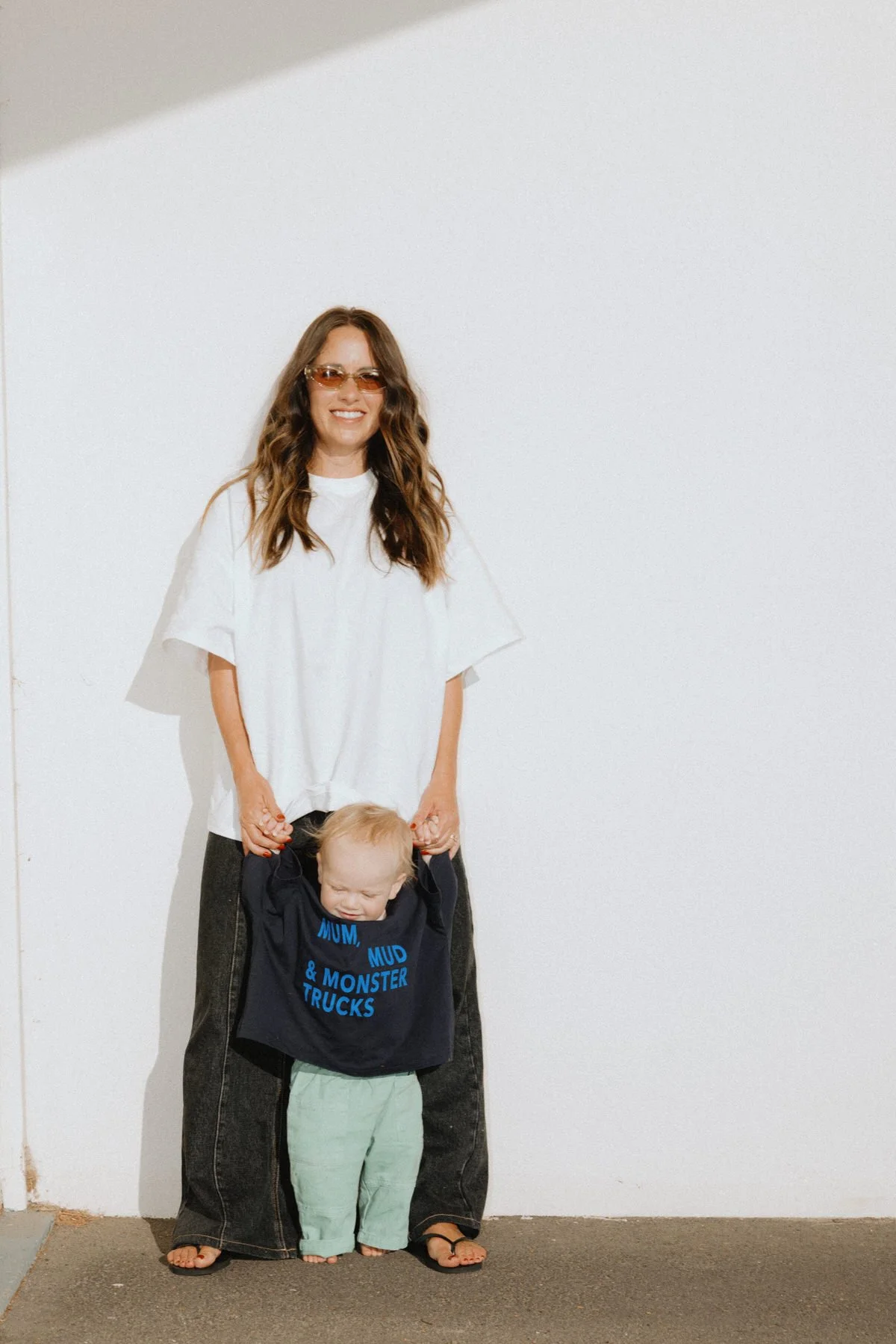 A woman with long, wavy brown hair wearing sunglasses, a white oversized T-shirt, and wide-leg jeans, smiling while holding the hands of a young boy with short blonde hair, who is wearing a navy blue sweatshirt with blue text about trucks and light-c