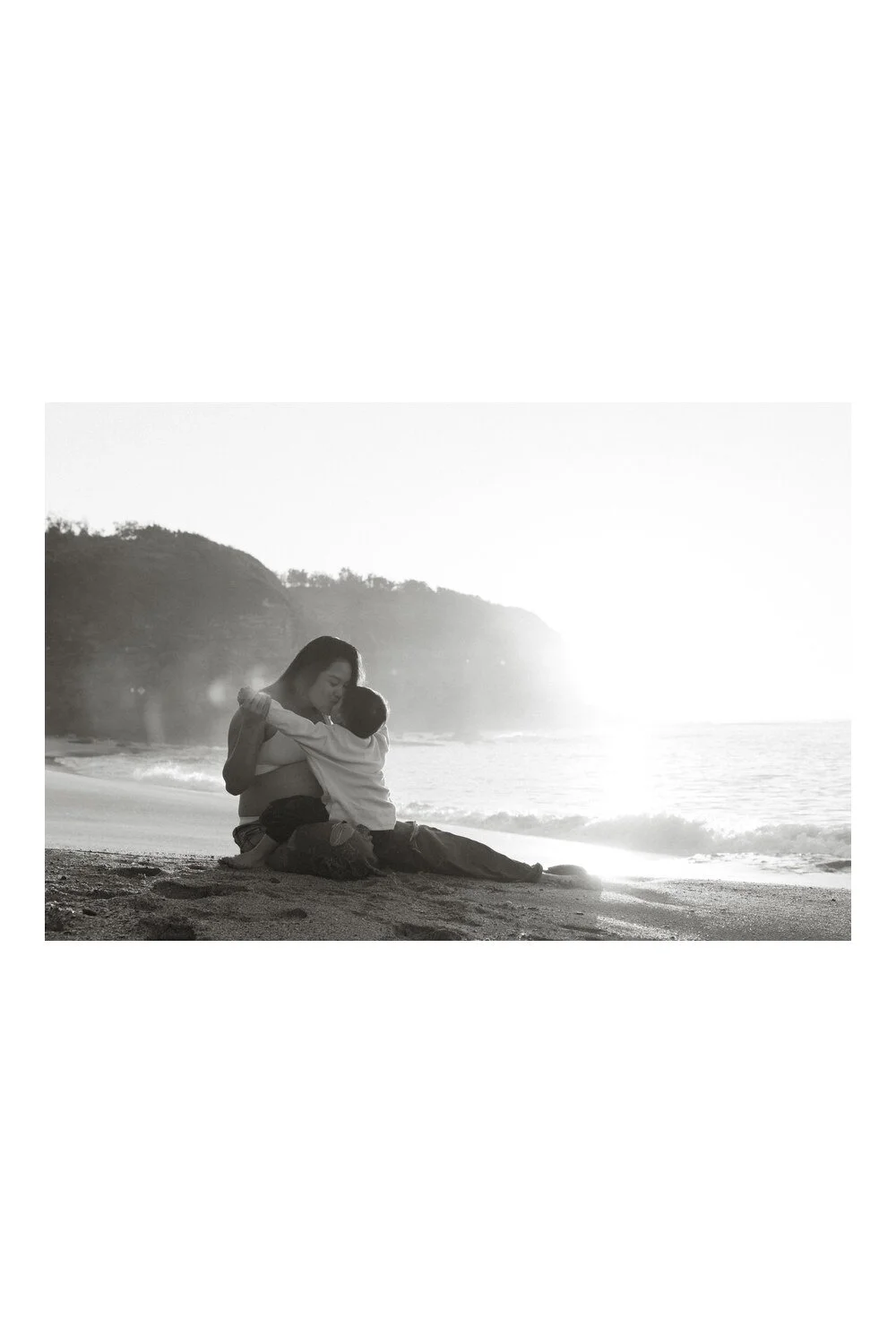 Black-and-white photograph of a mother sitting on the sand, cuddling her toddler beside the sea.