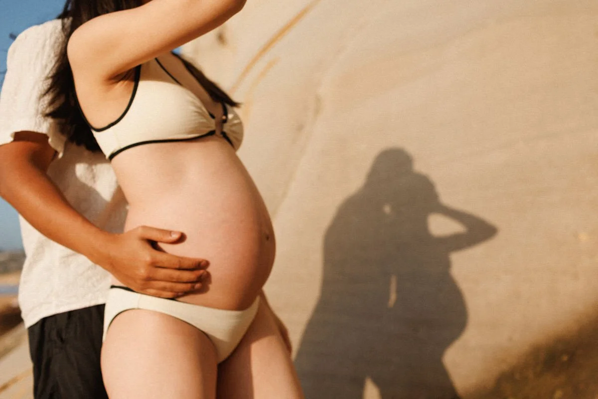A pregnant woman wearing a cream-colored lingerie set with black trim stands against a wall, with her hand resting on her belly. Someone behind her has their hand on her waist. The woman’s shadow is cast on the wall, showing her profile and her partner’s silhouette.