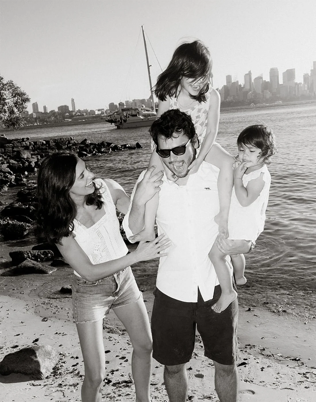 A black and white photo of a family at the beach. A man with sunglasses is carrying a young girl on his shoulders, and a woman is holding his arm while another young girl is standing next to him, touching his arm. They are all smiling and enjoying the moment with city skyline and boats in the background.