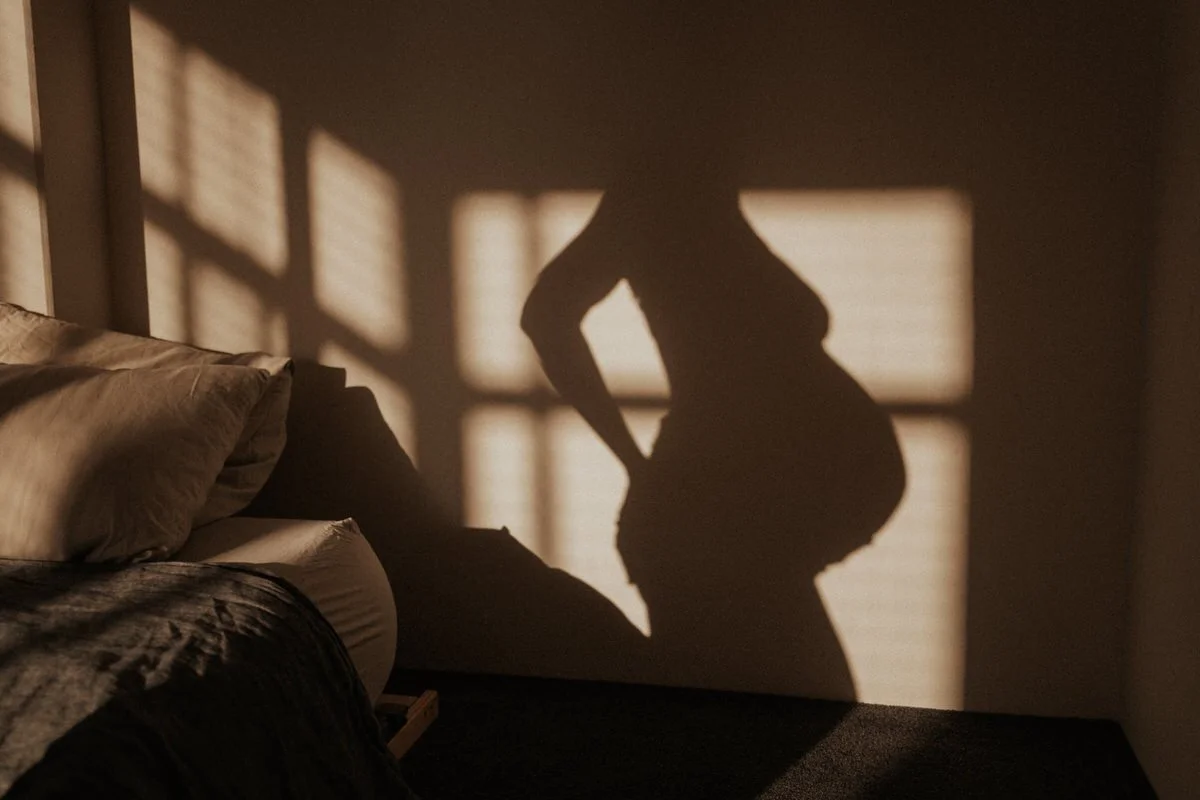 Silhouette of a pregnant woman standing with her hand on her hip in front of a window casting a shadow on the wall.