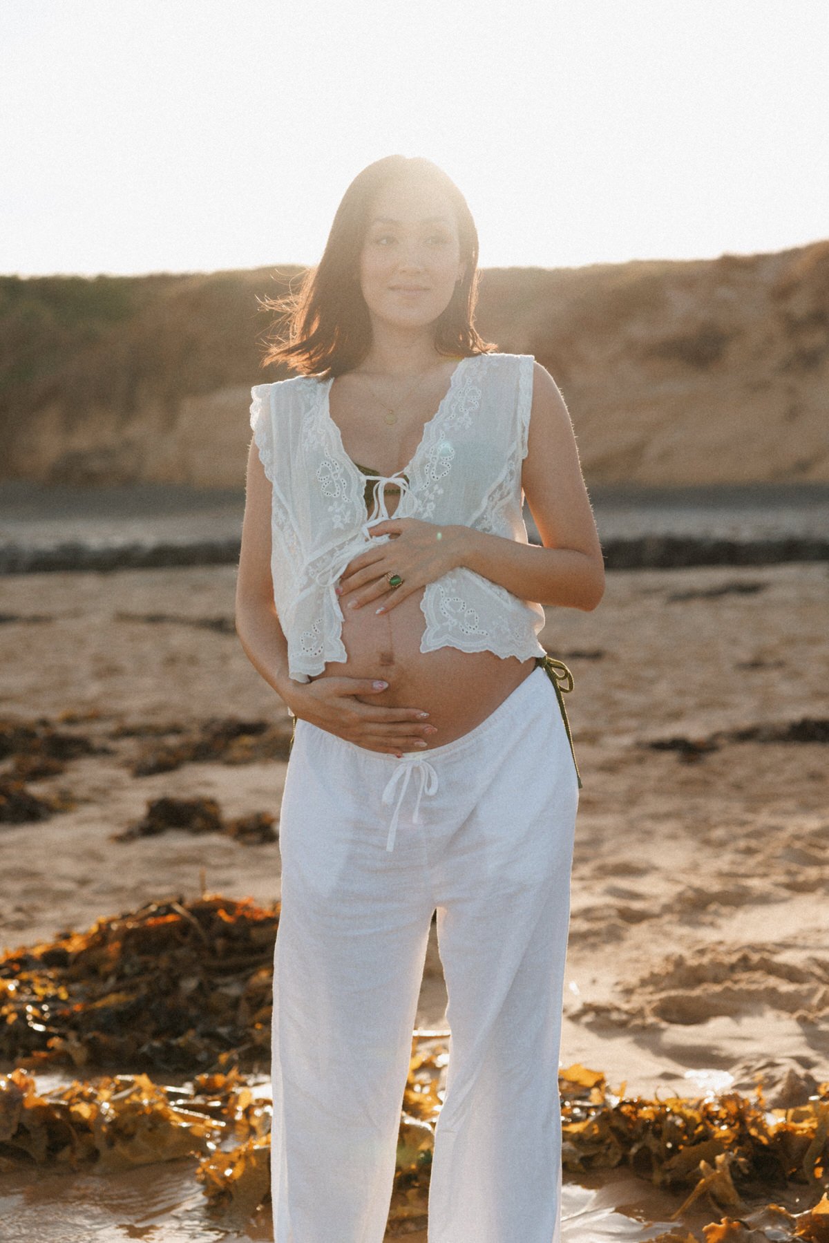 A pregnant woman with shoulder-length dark hair standing on a beach at sunset, wearing a white lace top and matching white pants, gently holding her baby bump with a smile.