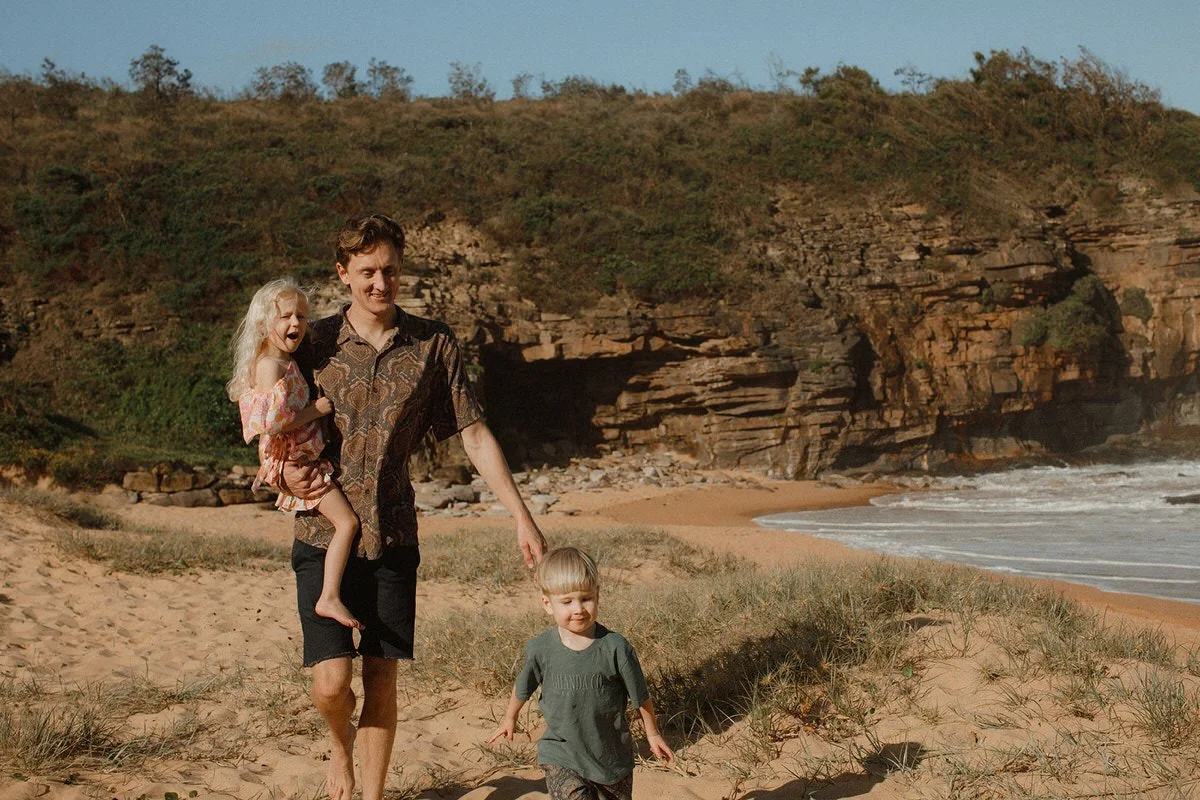 A man walking on a sandy beach with two young children, one girl and one boy, near rocky cliffs under a clear sky.