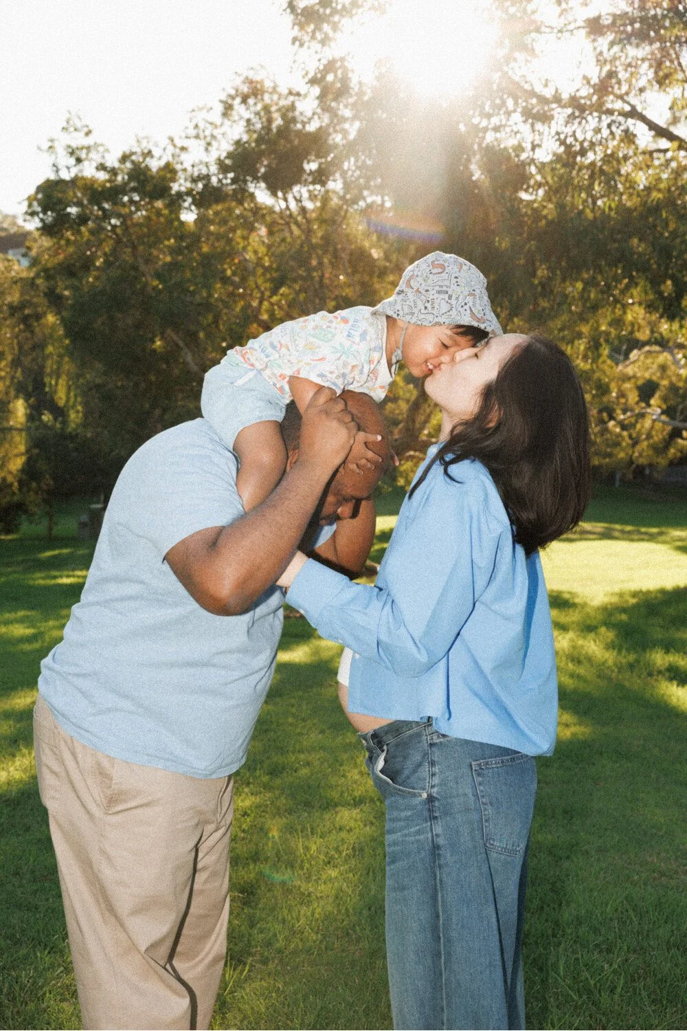 Family kiss: Parents kissing their toddler during a sunlit maternity session in the park.