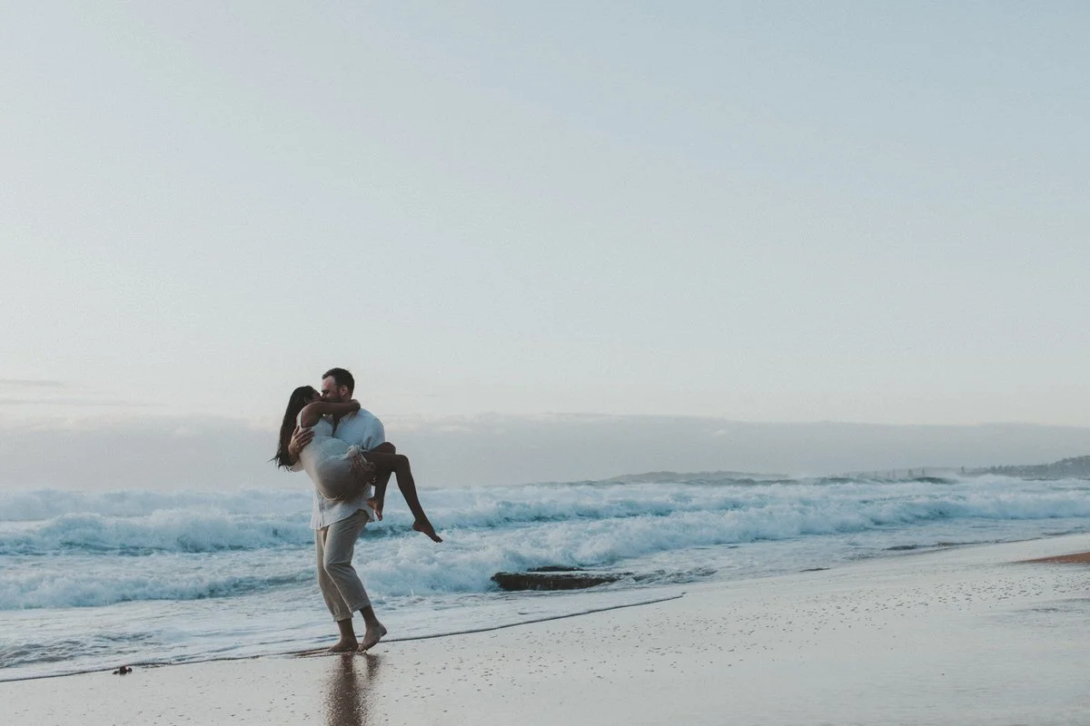 A man carrying a woman in his arms on the beach near the ocean, with waves in the background during dusk