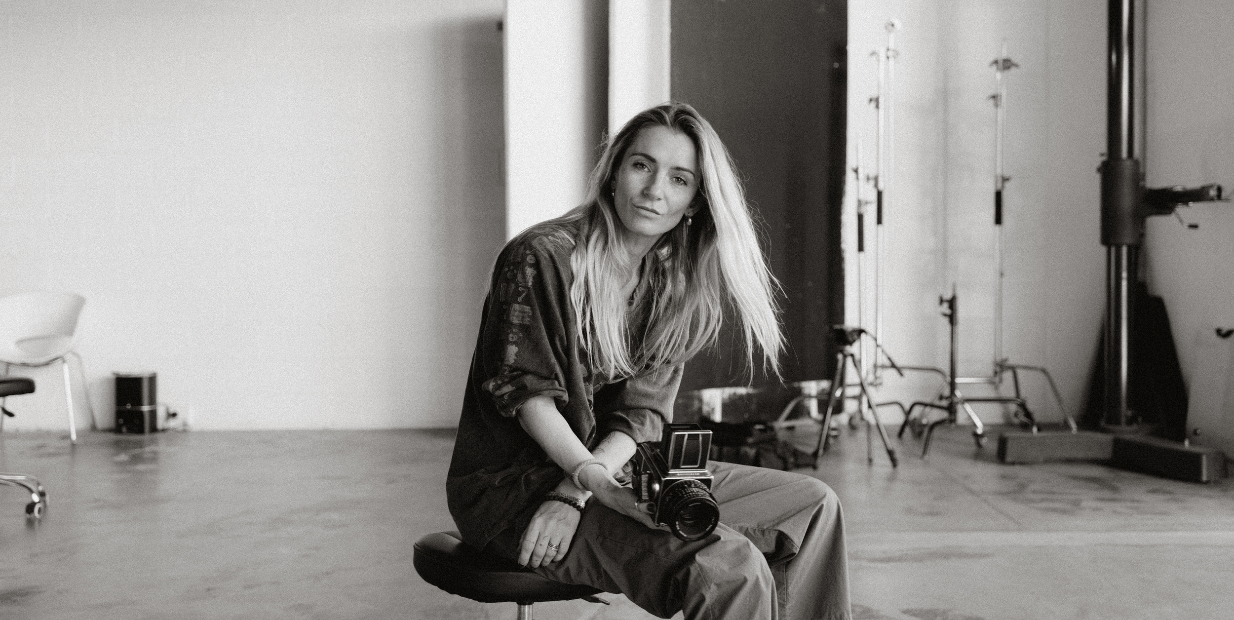 A woman sitting on a stool in a photography studio holding a camera, with studio equipment and lighting in the background, in black and white.