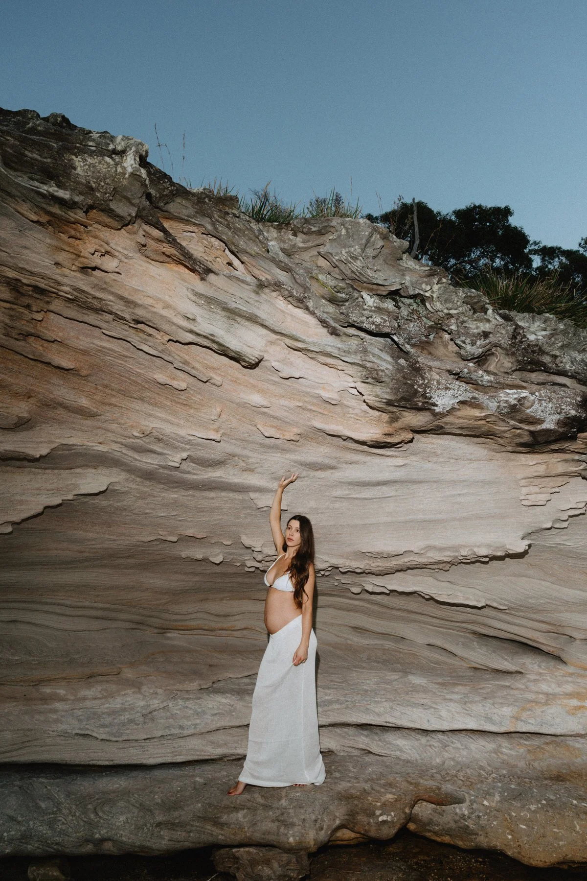 A woman in a white dress standing barefoot in a cave with layered, textured rock walls, raising one arm.