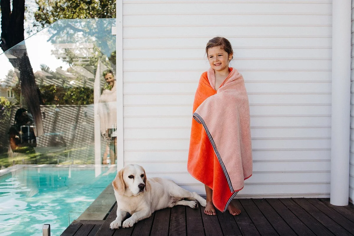 Young girl wrapped in a pink and orange towel standing on wooden deck next to a white dog lying down. In the background, there's a white house, a glass door with reflections, and a backyard pool.