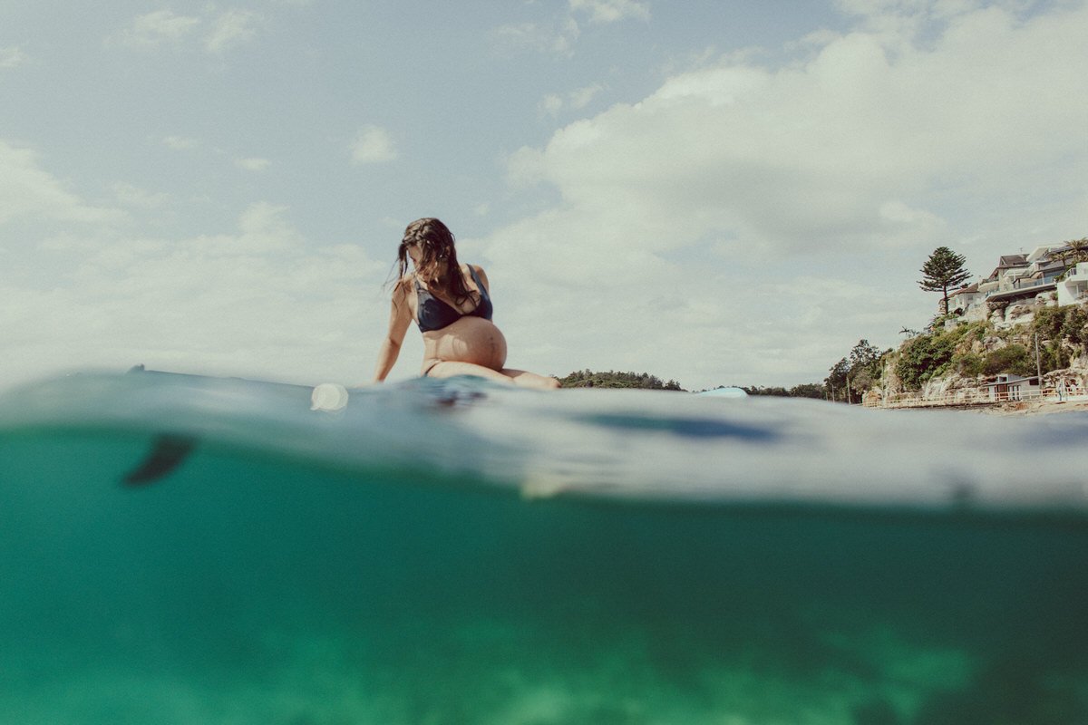 A pregnant woman in a black swimsuit sitting on a surfboard in the water with a shoreline of houses and trees in the background under a cloudy sky.