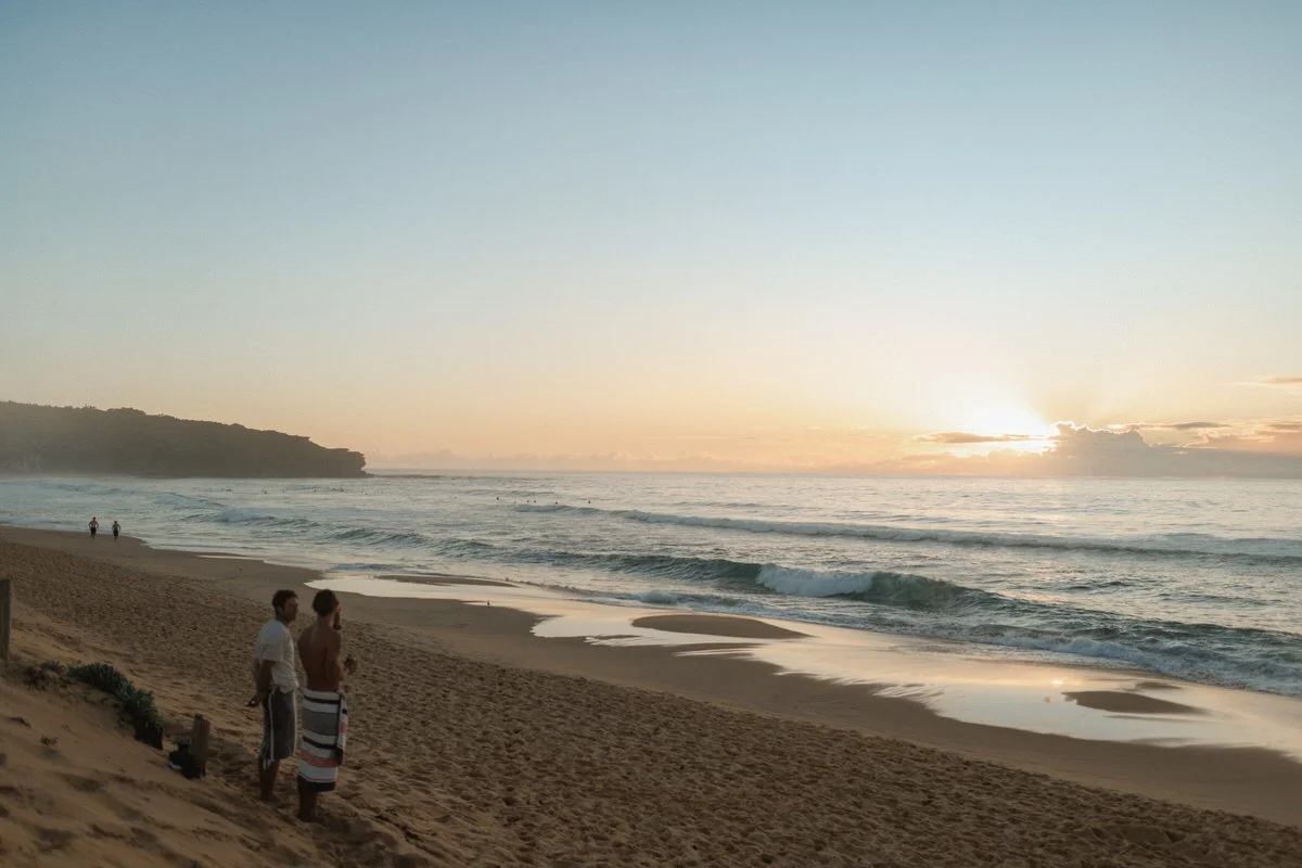 People standing on a sandy beach at sunset, with ocean waves and a distant landmass in the background.