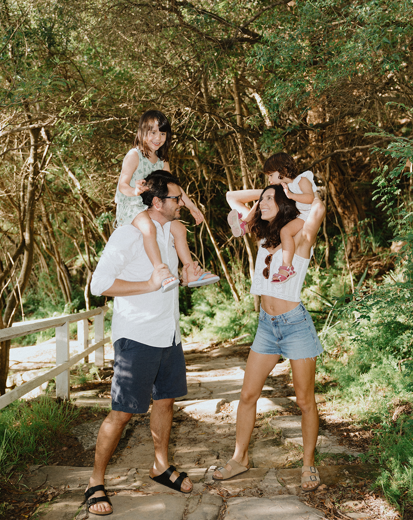 A family of four enjoying a walk through a wooded area, with the father and mother carrying their two young daughters on their shoulders.