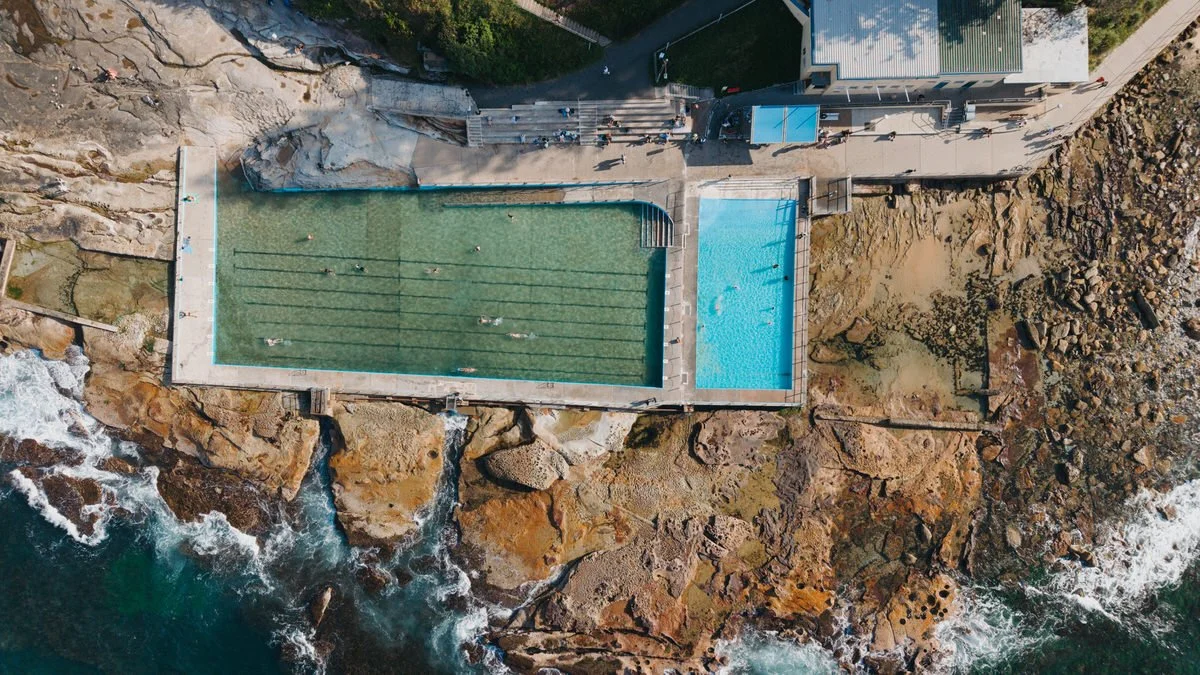 An aerial view of an outdoor swimming pool situated on a rocky shoreline, with a smaller pool nearby and a concrete pathway leading to a building.
