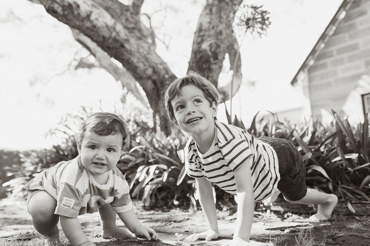 Two young boys are doing push-ups outdoors on a dirt ground with plants and a large tree behind them, one boy smiling and the other looking at the camera.